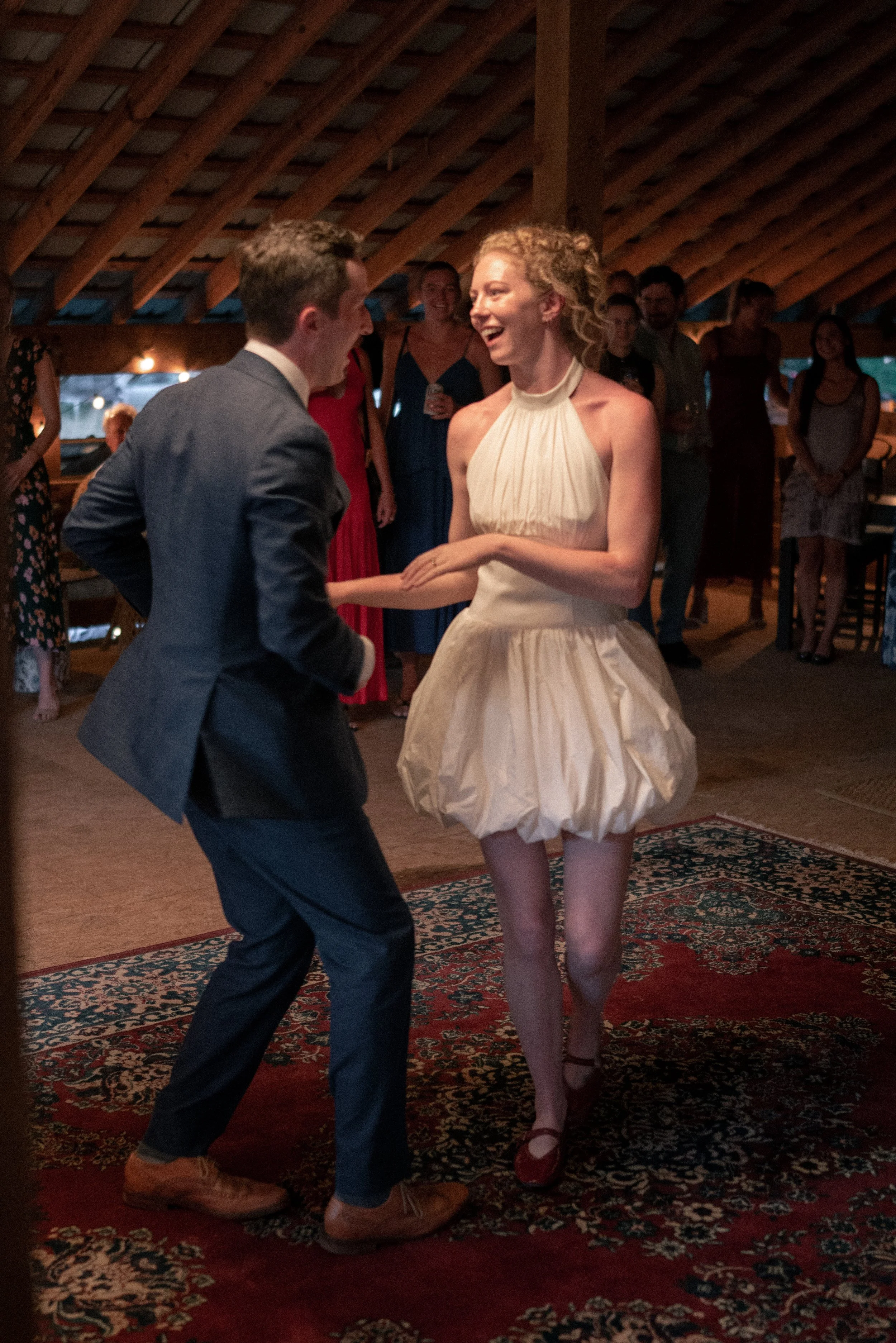 A man and woman dancing together at a social gathering indoors, with other people watching and smiling in the background.