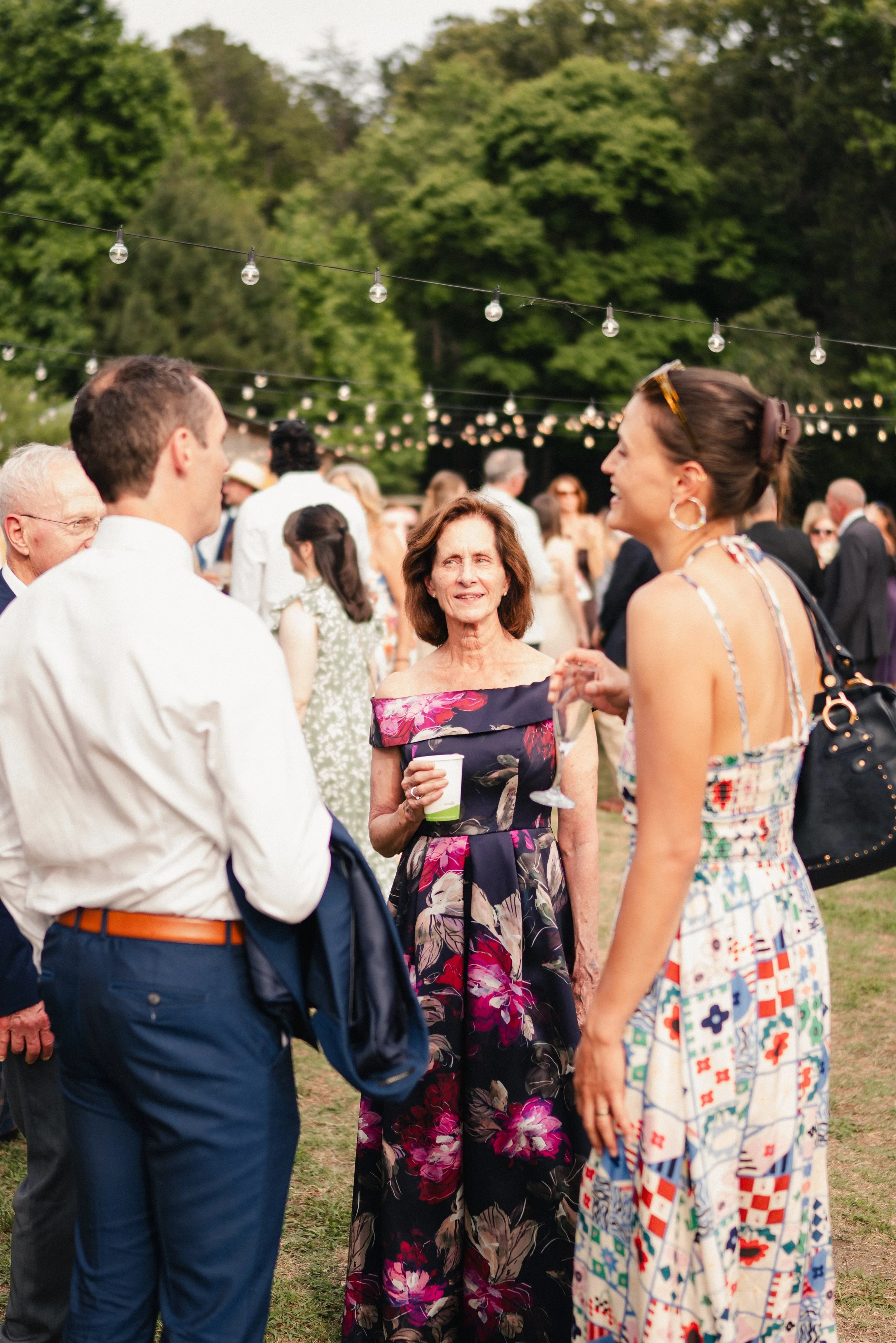 People socializing outdoors during daytime at a gathering with string lights overhead, amidst green trees.