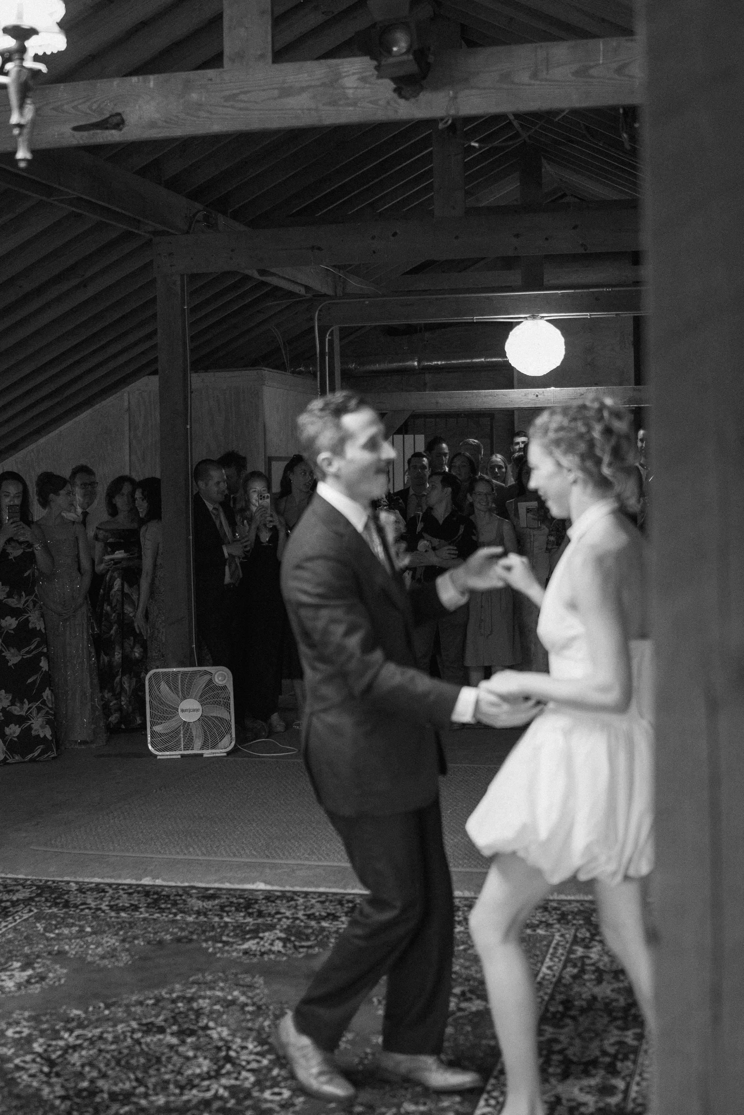 A couple dances together in the foreground while a crowd of guests watches in the background at an indoor wedding reception with wooden beams and a hanging paper lantern.