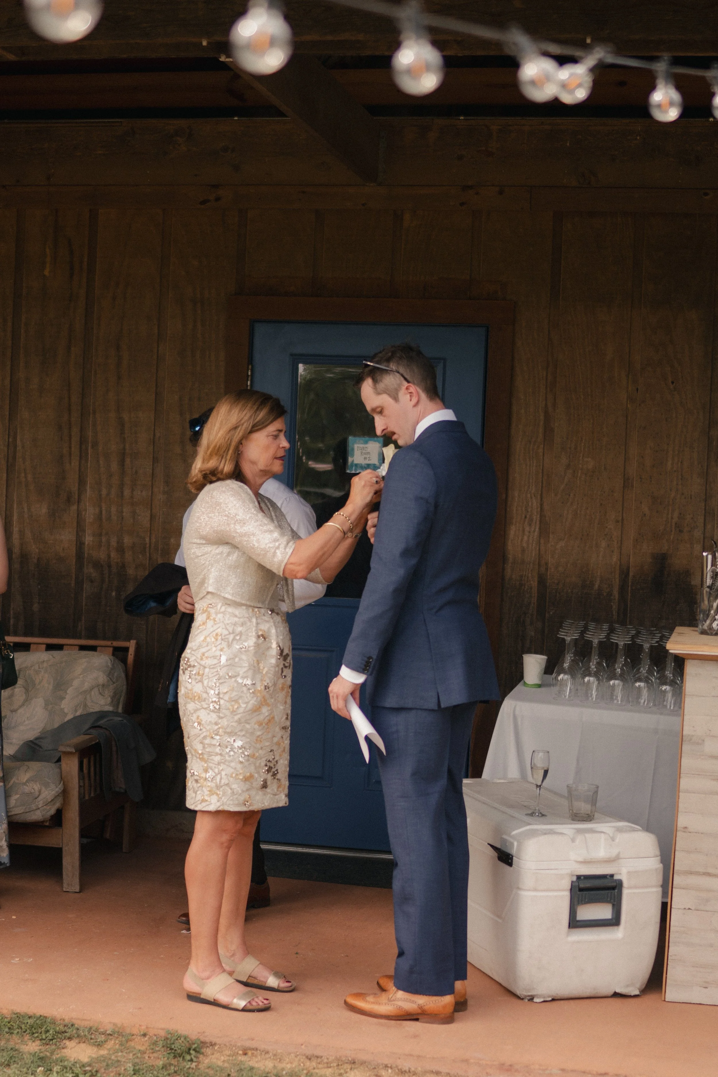 A woman in a light-colored dress is being pinned with a flower corsage by a young man in a blue suit at an indoor event. There is a table with glasses and a cooler nearby.