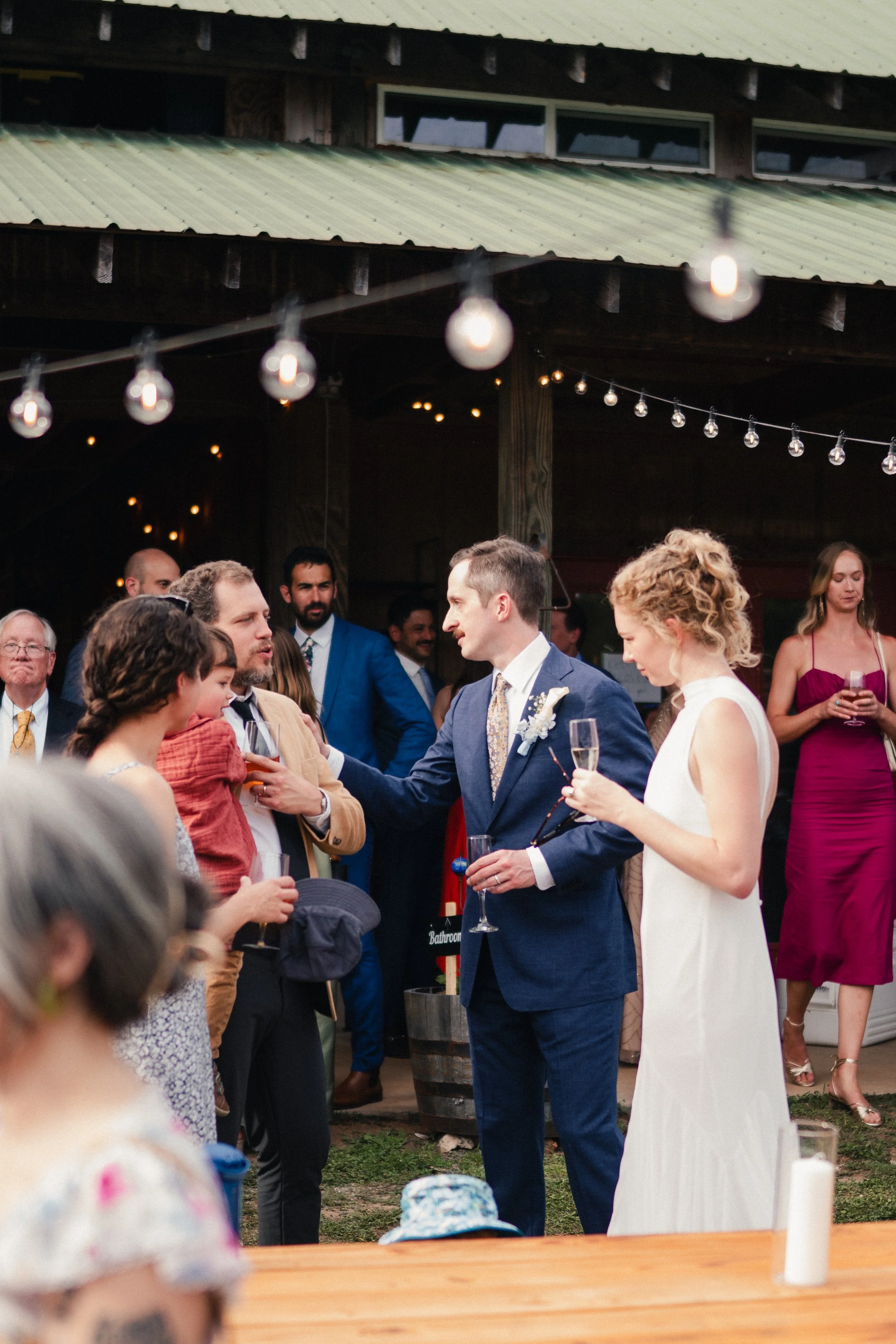People gathered outdoors at a wedding reception, with some holding drinks, under string lights near a rustic building with a green metal roof.