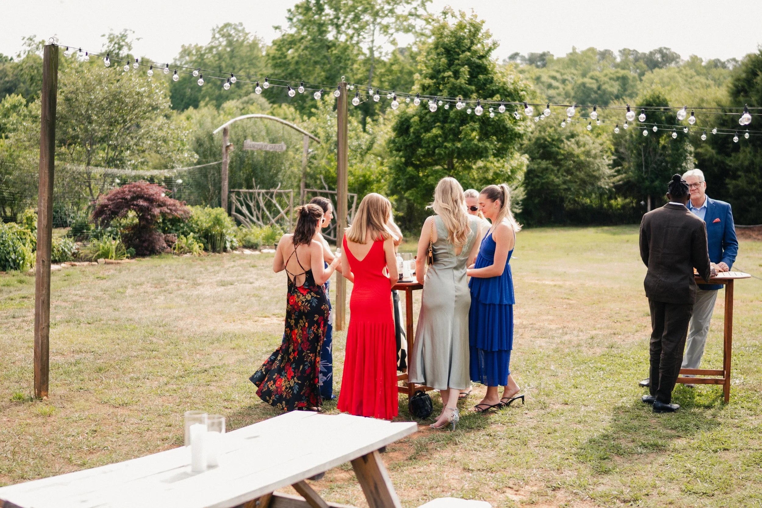 Group of people dressed in formal attire standing outdoors under string lights in a garden setting, engaged in conversation.