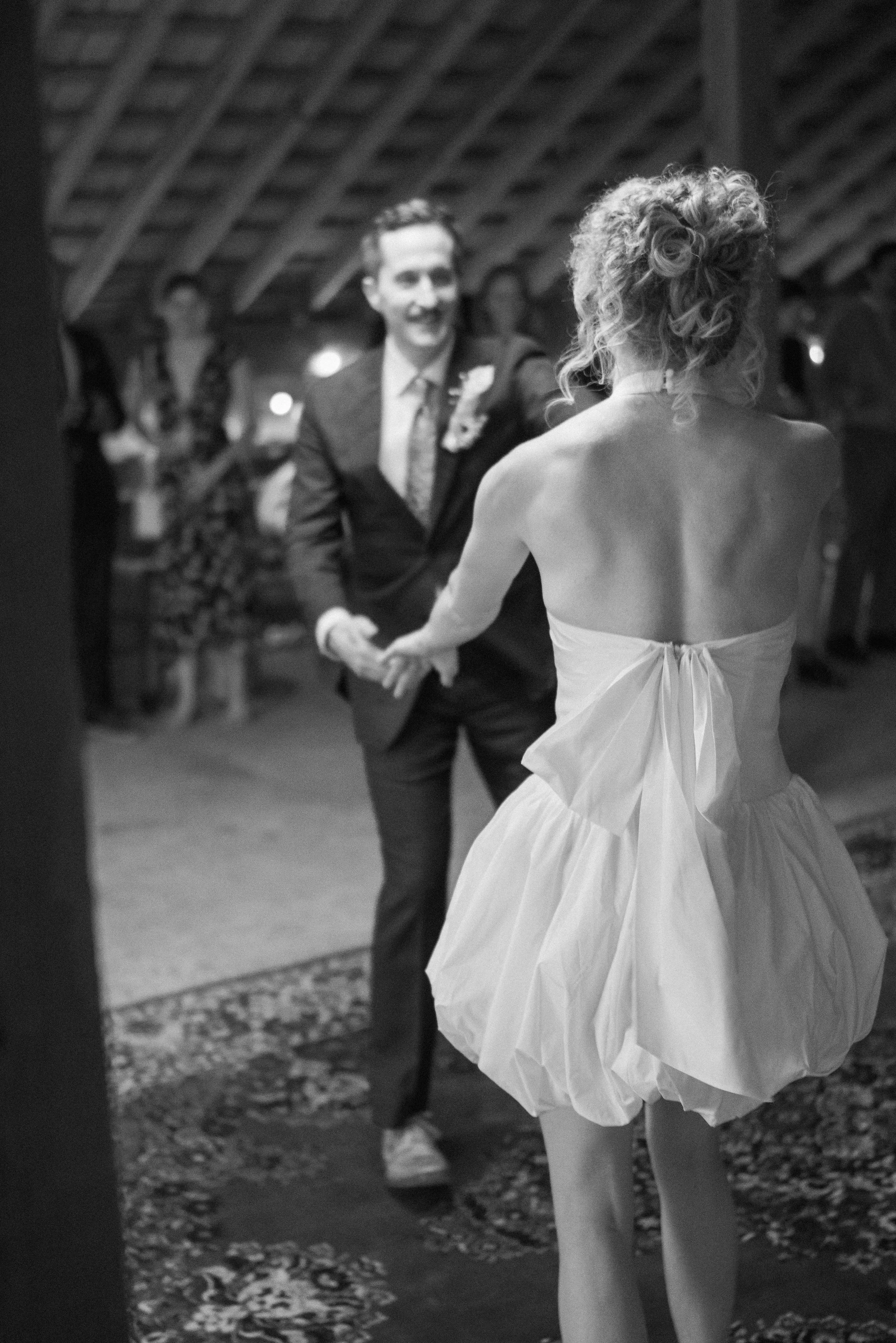 A woman in a strapless dress and ballet tights dancing with a man in a tuxedo at a celebration or wedding reception, surrounded by guests in a rustic venue with a wooden ceiling.