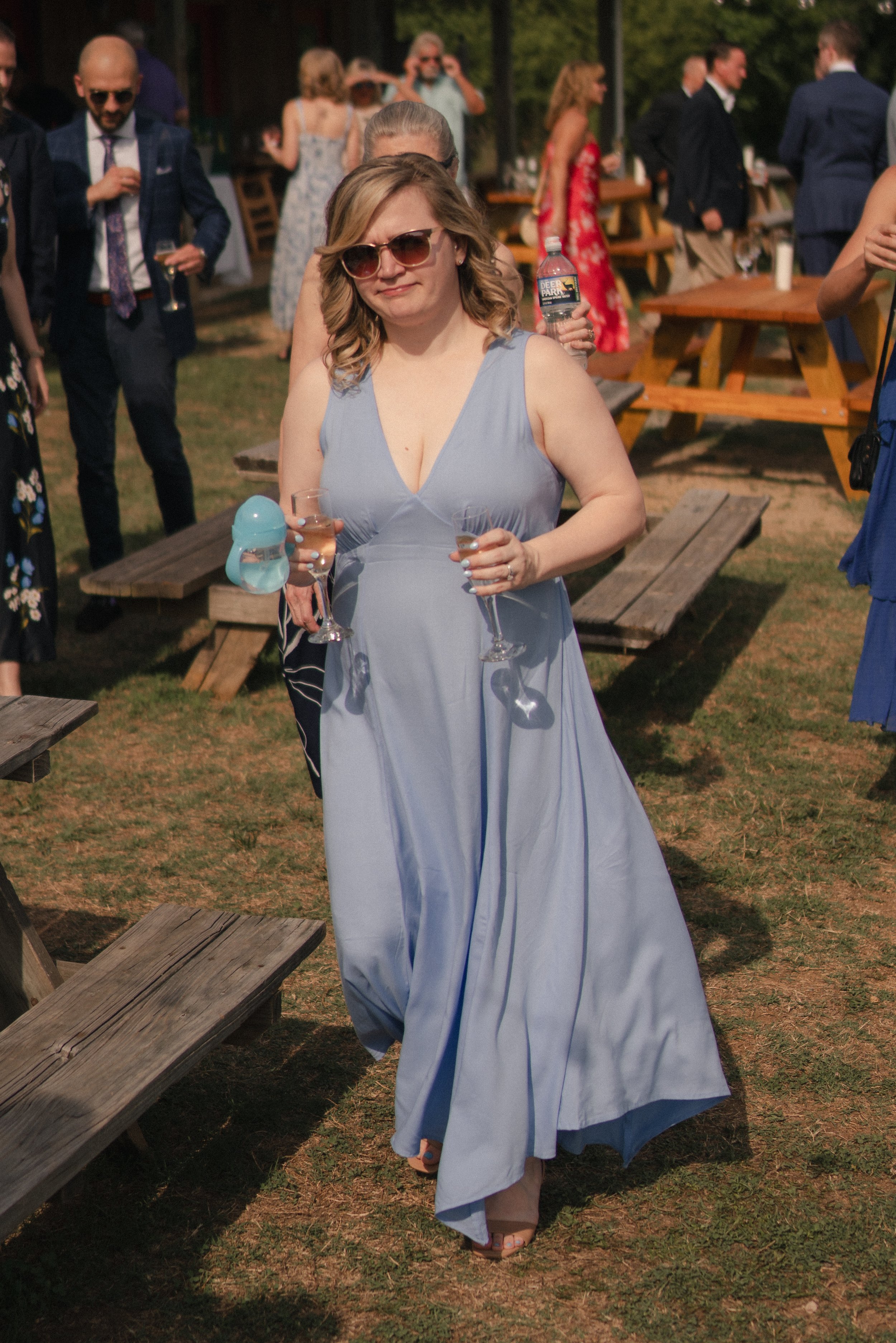 A woman in a blue dress and sunglasses holding a drink and a water bottle at an outdoor gathering.