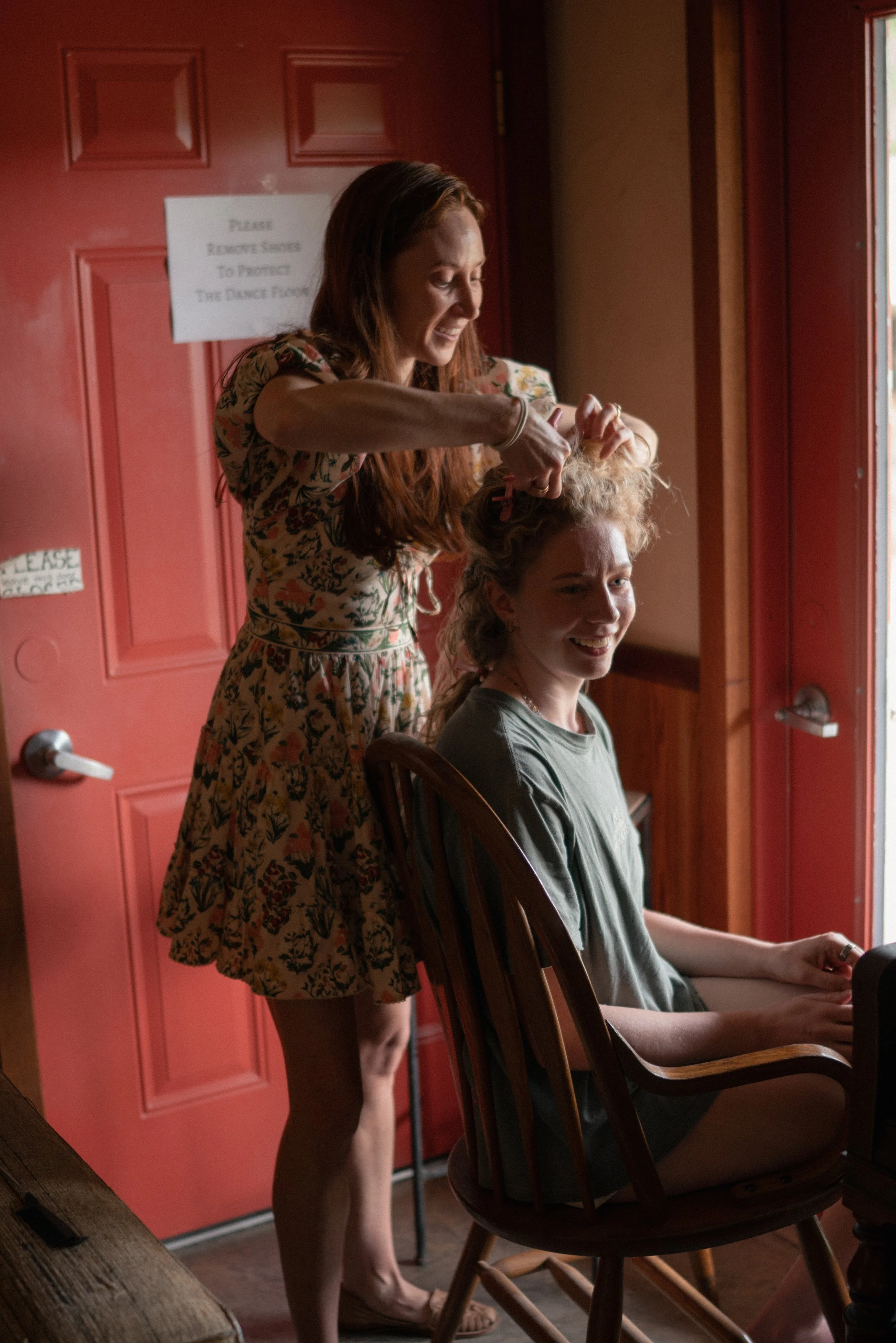 A woman with red hair styling another woman's blonde hair inside a room with red walls and a window. The woman sitting is smiling and holding a phone, while the other woman, standing, is focused on her task.