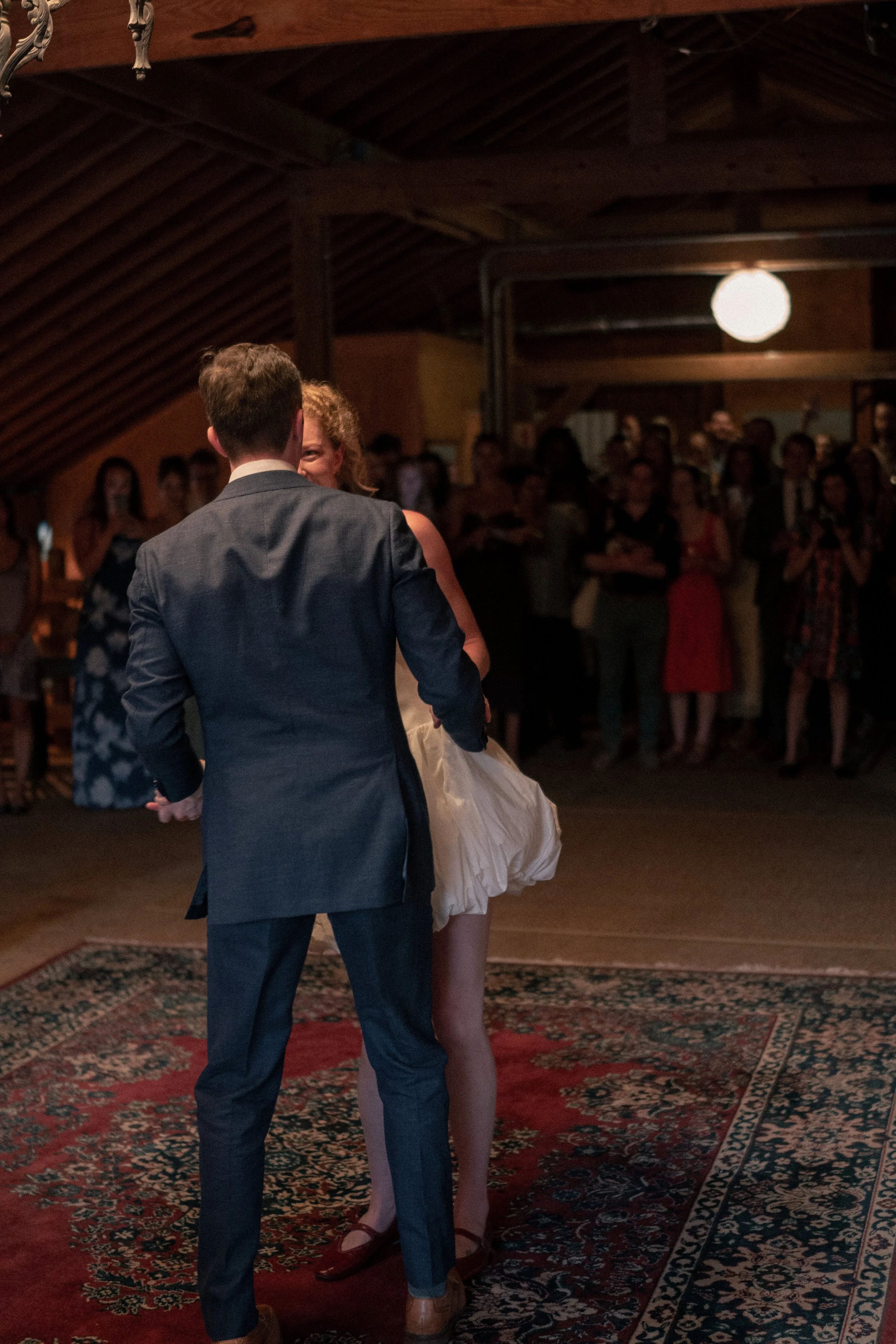 A couple is dancing closely in a dimly lit venue with a wooden ceiling, and an audience of onlookers watching them. The man is wearing a dark suit, and the woman has curly hair and is dressed in a white dress. The group in the background includes wom