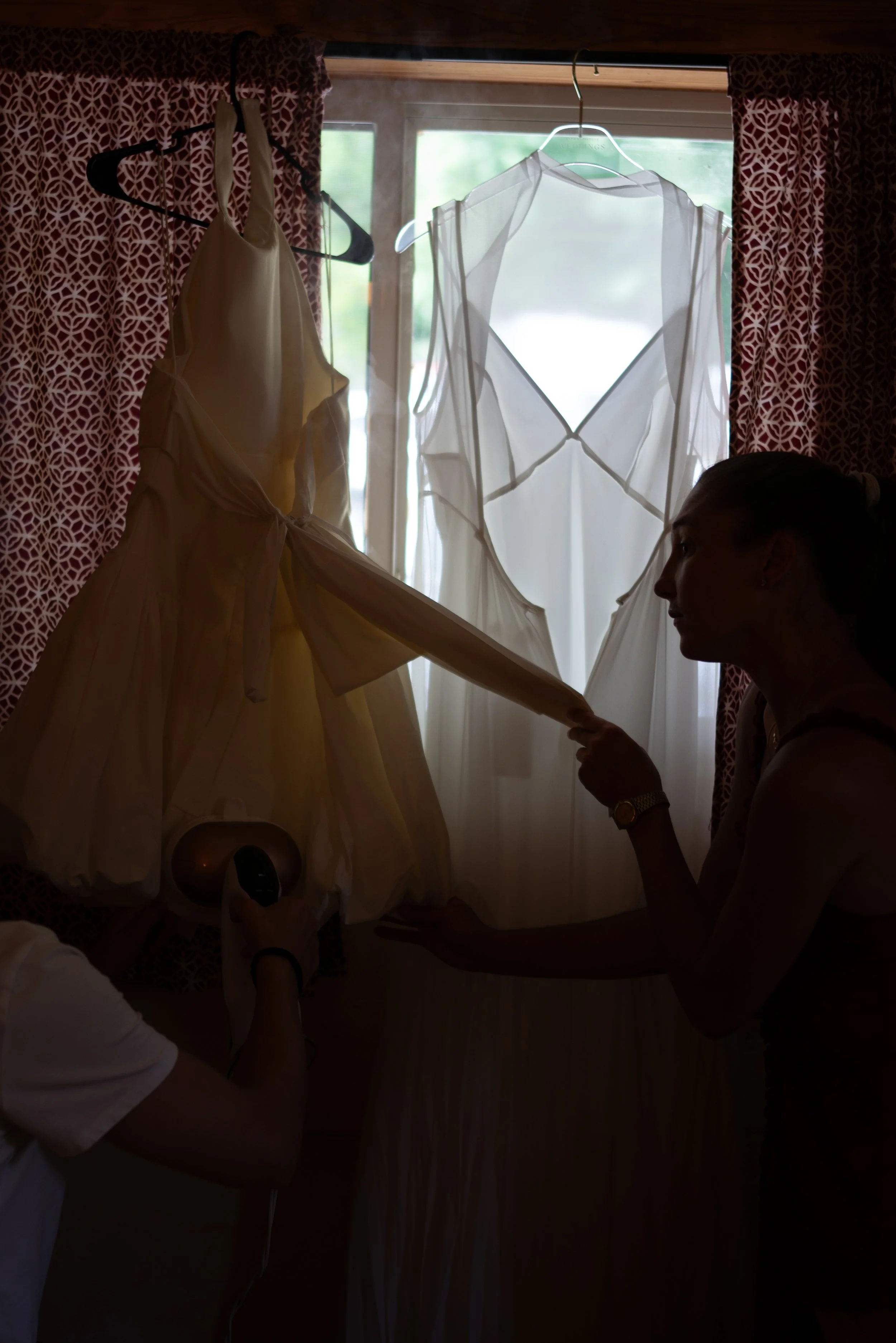 People hanging wedding dresses and adjusting a white wedding gown near a window with red patterned curtains.