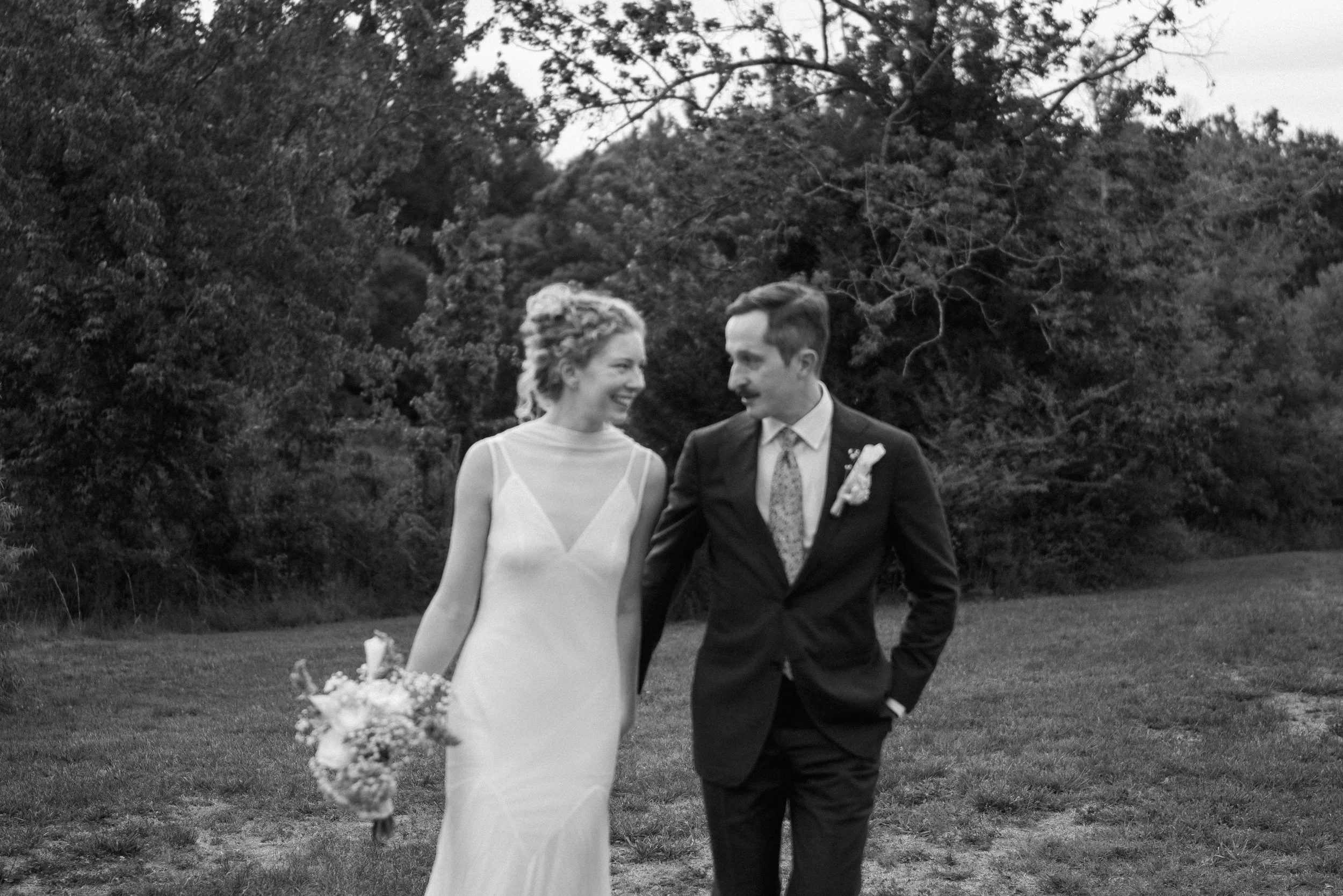 A black-and-white photo of a smiling bride and groom walking outdoors on their wedding day, holding hands, with trees in the background.
