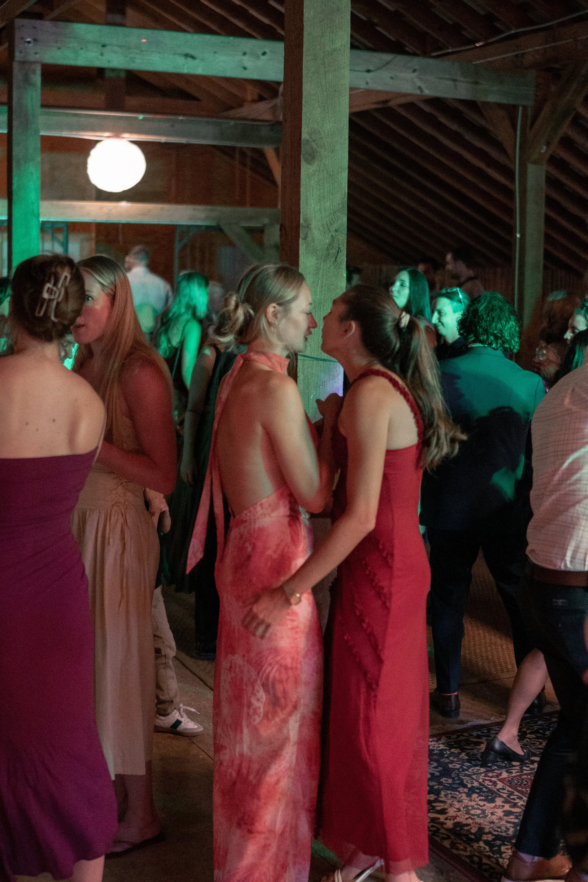 Group of people socializing at an indoor event, some wearing formal attire, in a rustic wooden space with a high ceiling and decorative rug.