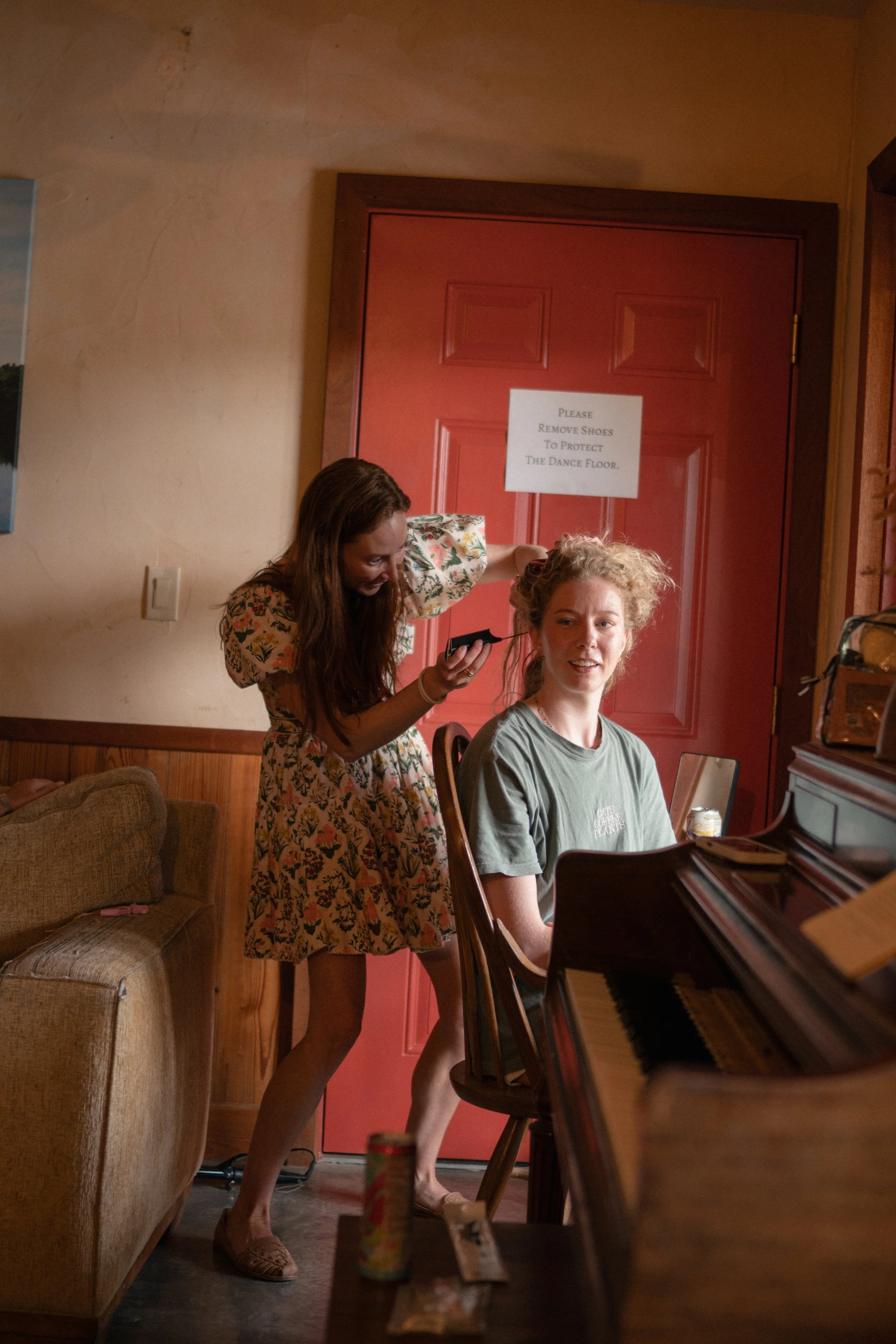 A woman sitting at a piano with her hair in a messy bun, wearing a green t-shirt. Another woman, standing, is styling her hair. They are in a cozy room with a red door and a sign that says, "Please Remove Shoes To Protect The Dance Floor."