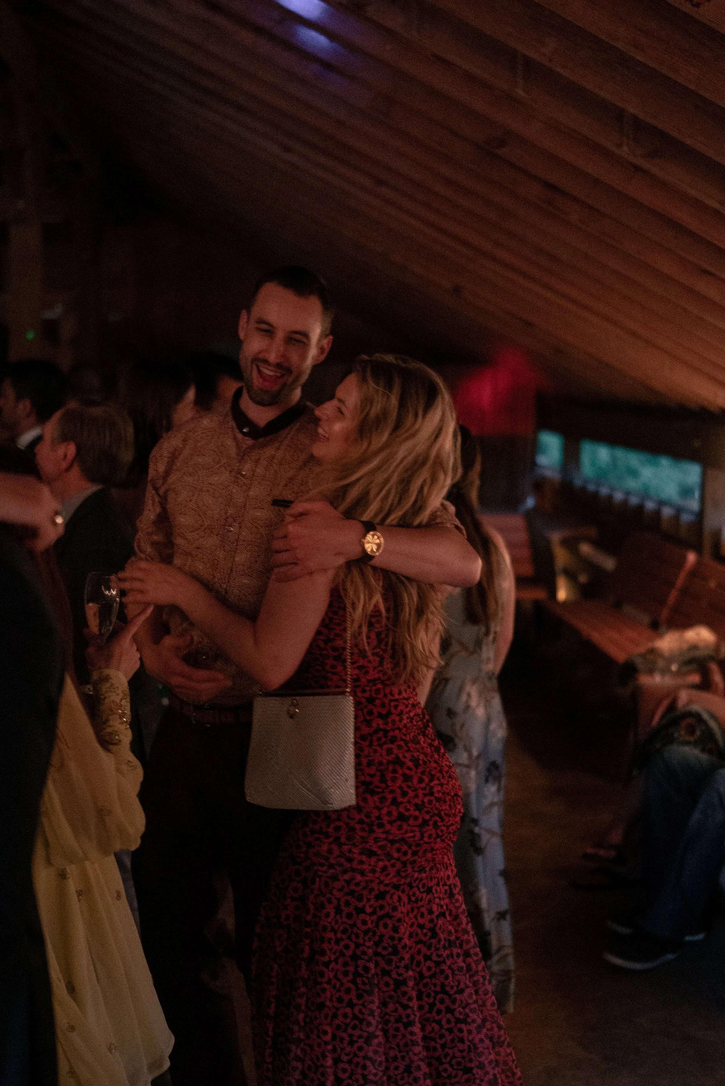 A group of people at a social event, with a focus on a man and woman hugging and smiling, in a warmly lit indoor setting with a wooden ceiling.