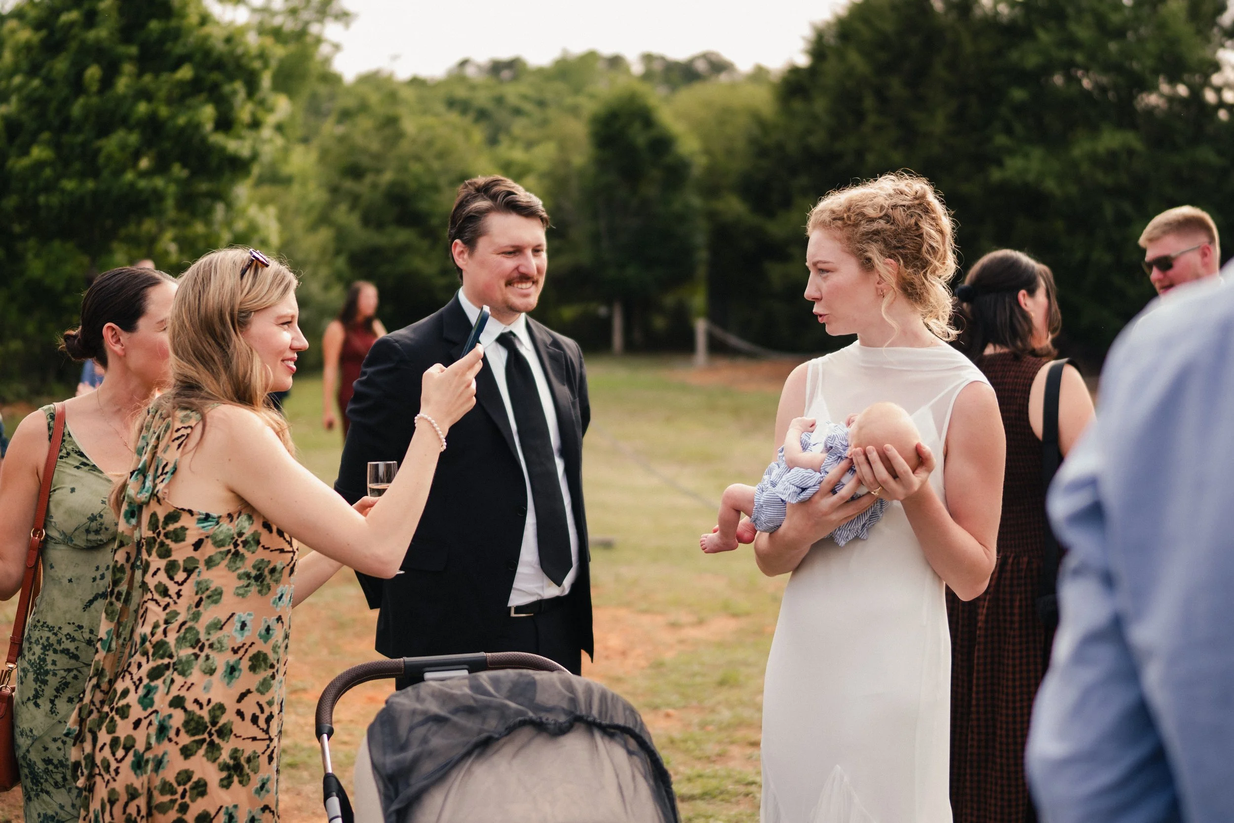A group of people at an outdoor gathering, with a woman holding a baby, others socializing, and a man in a suit smiling, set against a green, wooded background.