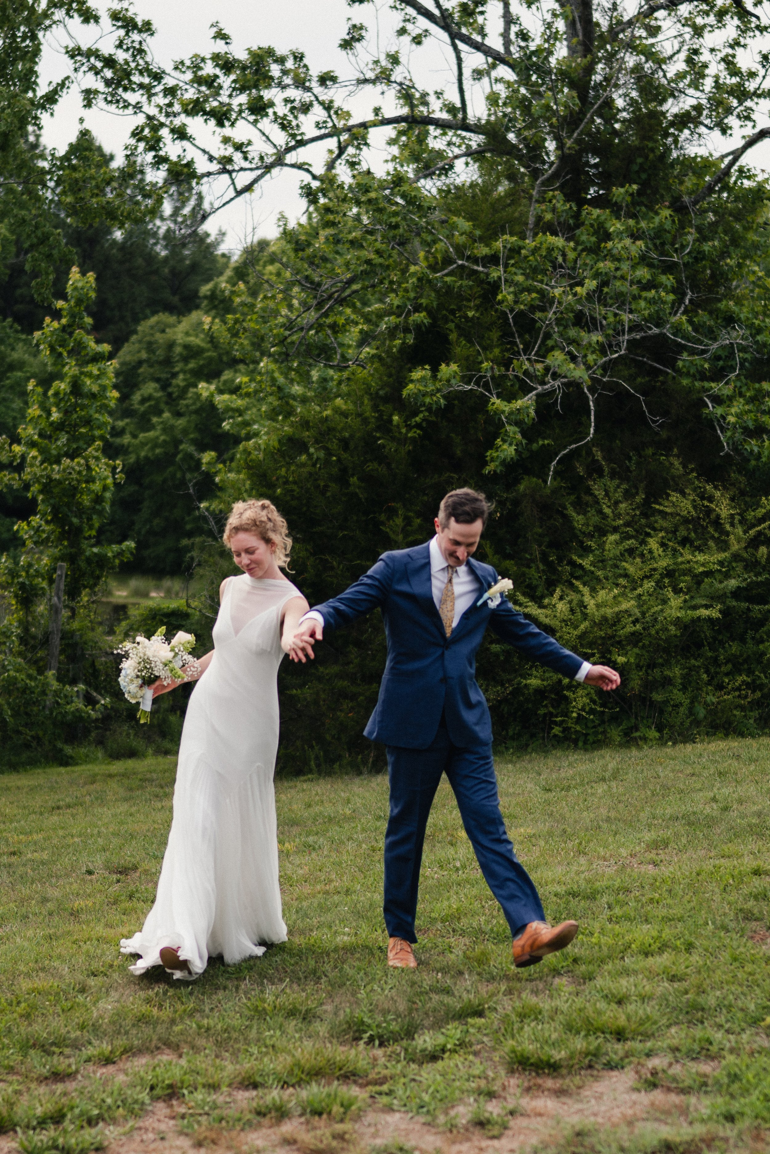 A bride and groom dancing outdoors on a grassy field during their wedding celebration, with trees in the background.