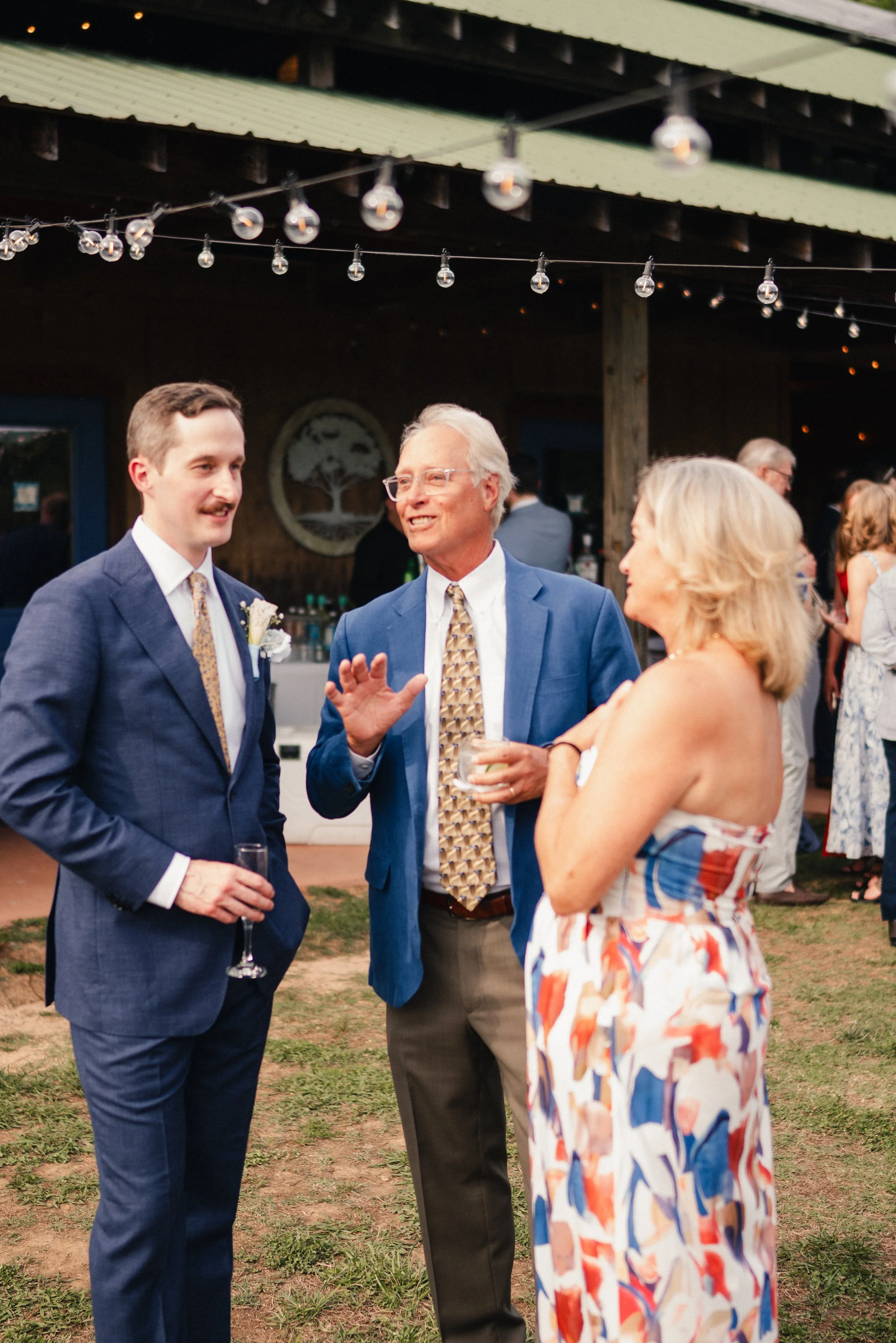 Three people at an outdoor event, a young man in a navy suit with a white shirt and tie, an older man in a blue suit and patterned tie, and a woman in a colorful dress, engaged in a conversation under string lights at dusk.