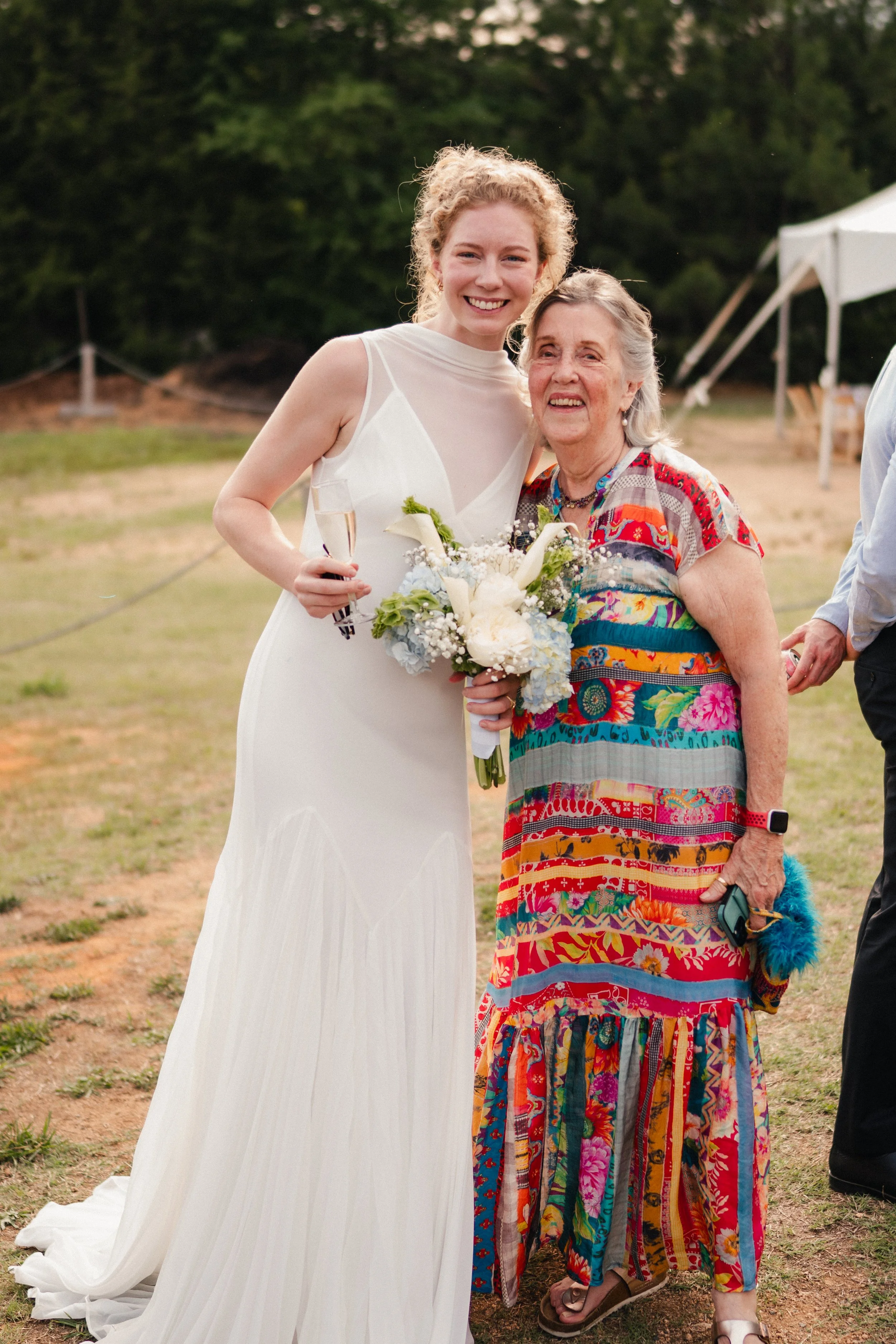 A young woman in a white dress holding a champagne glass and a bouquet, smiling with an elderly woman in colorful, patterned dress and sandals in an outdoor setting with tents and trees in the background.
