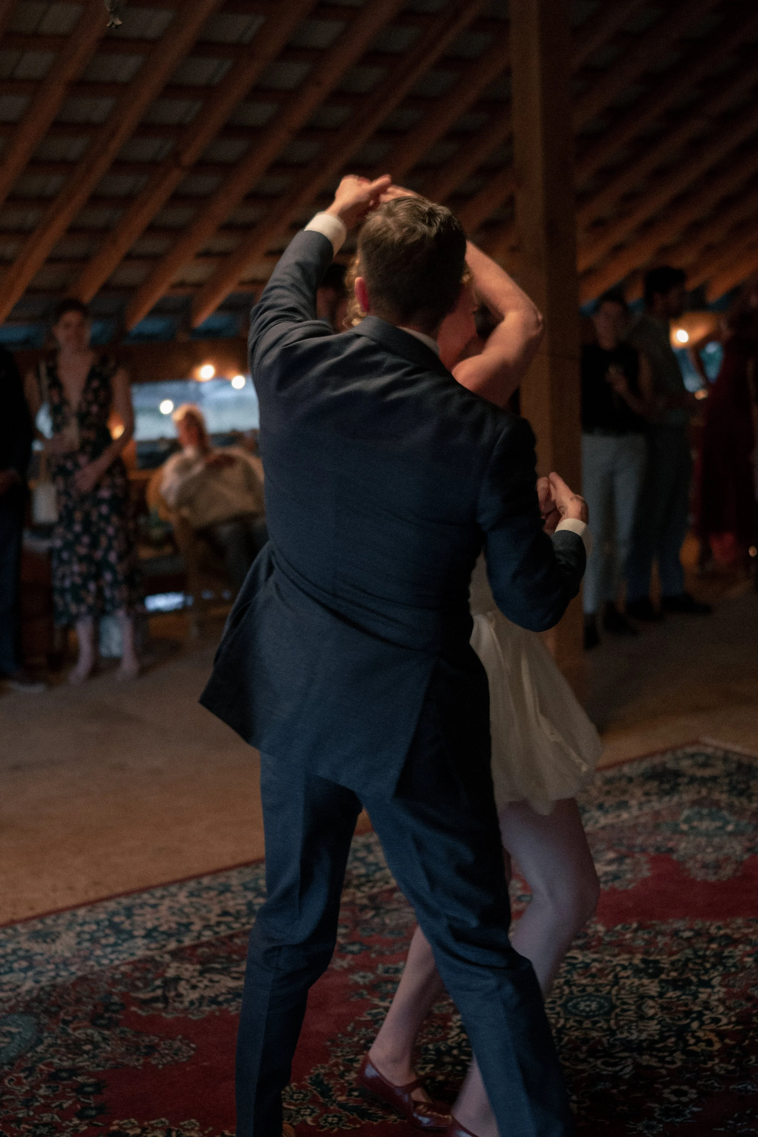 A couple dancing closely at an indoor event, with a patterned carpet and warm lighting, while other guests are visible in the background.