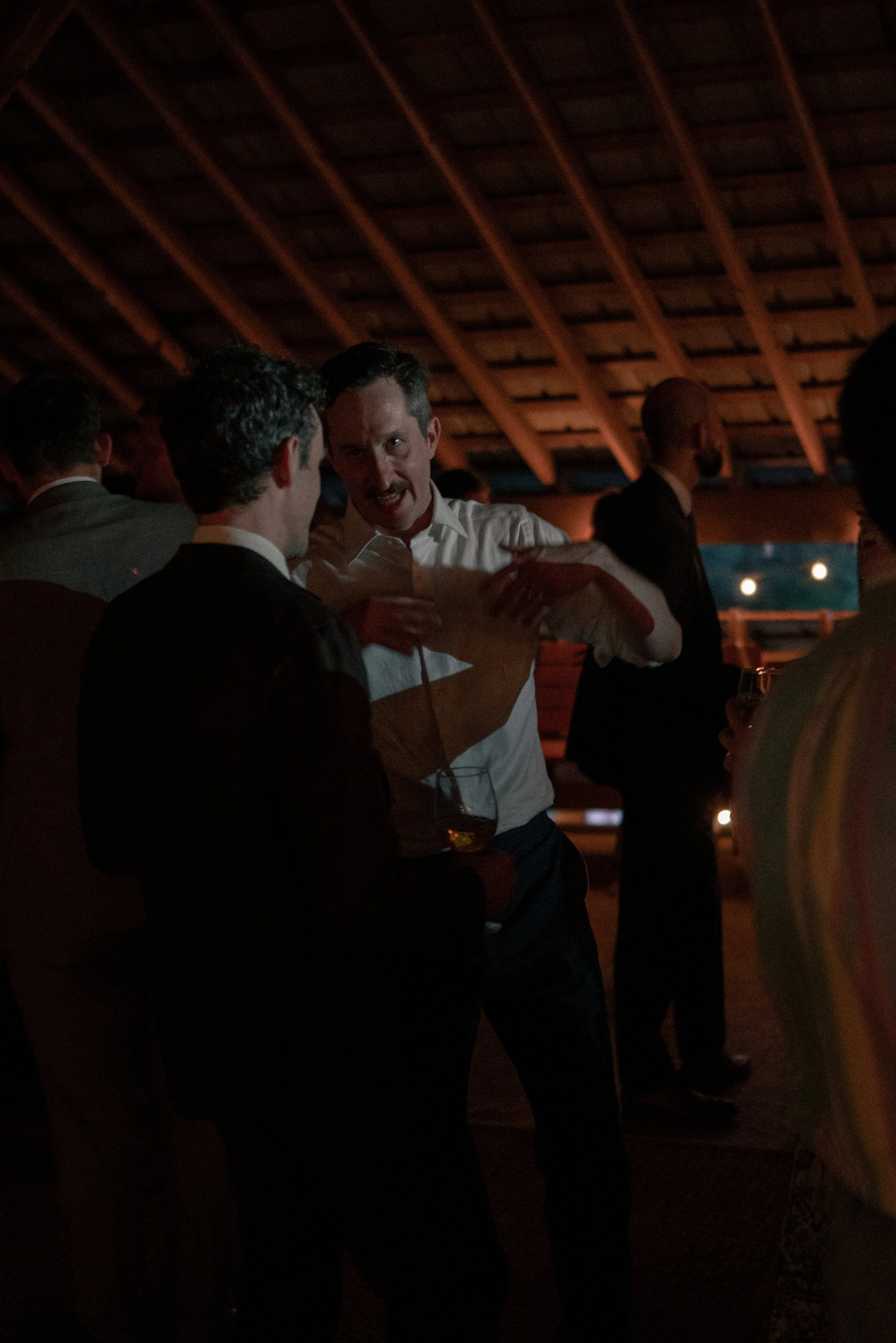 Men at a social gathering in a dimly lit venue, engaged in conversation and holding drinks, with wooden ceiling beams overhead.