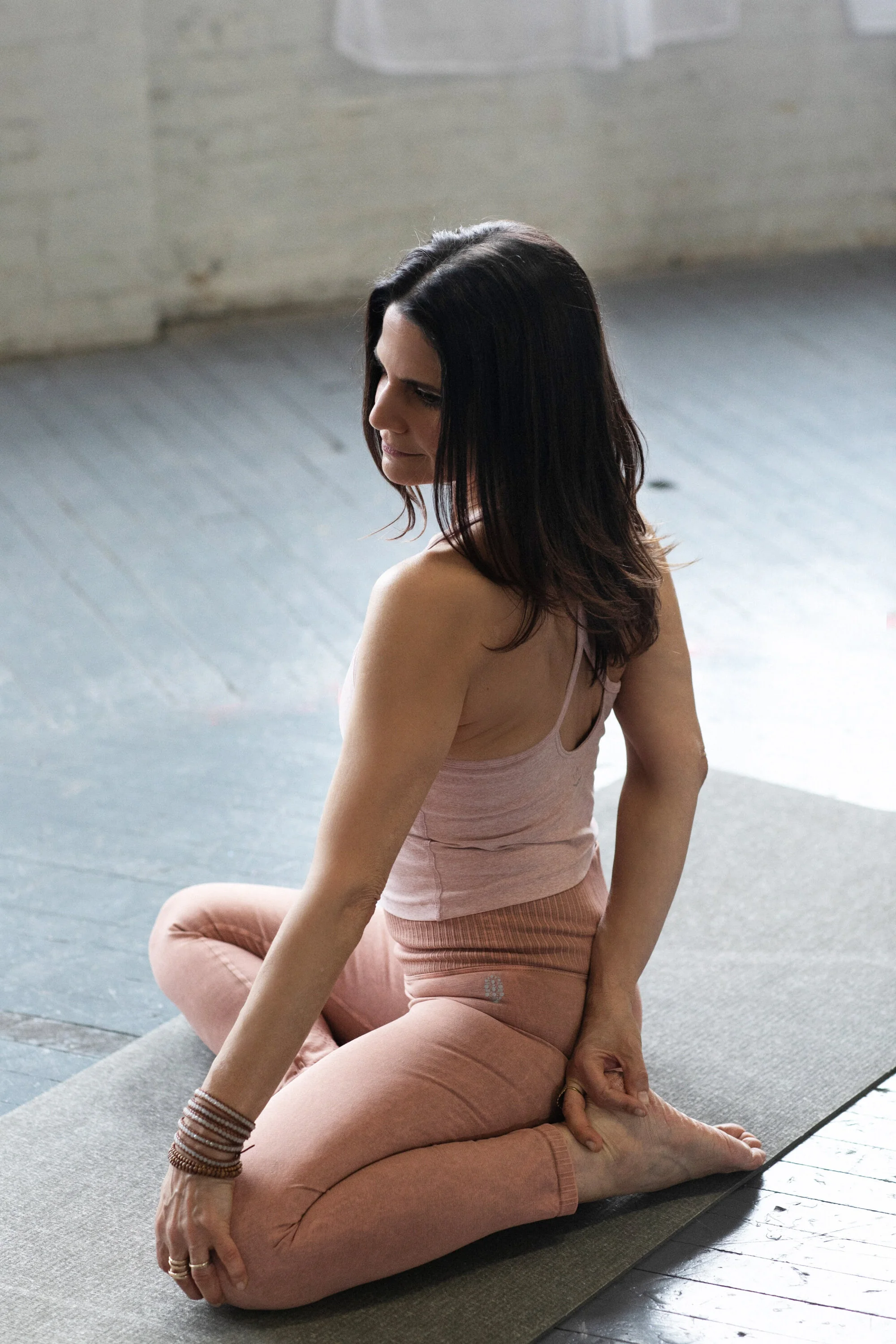 Woman practicing yoga on a mat, sitting in a cross-legged position with her left hand holding her right ankle, inside a room with wooden floors and brick walls.