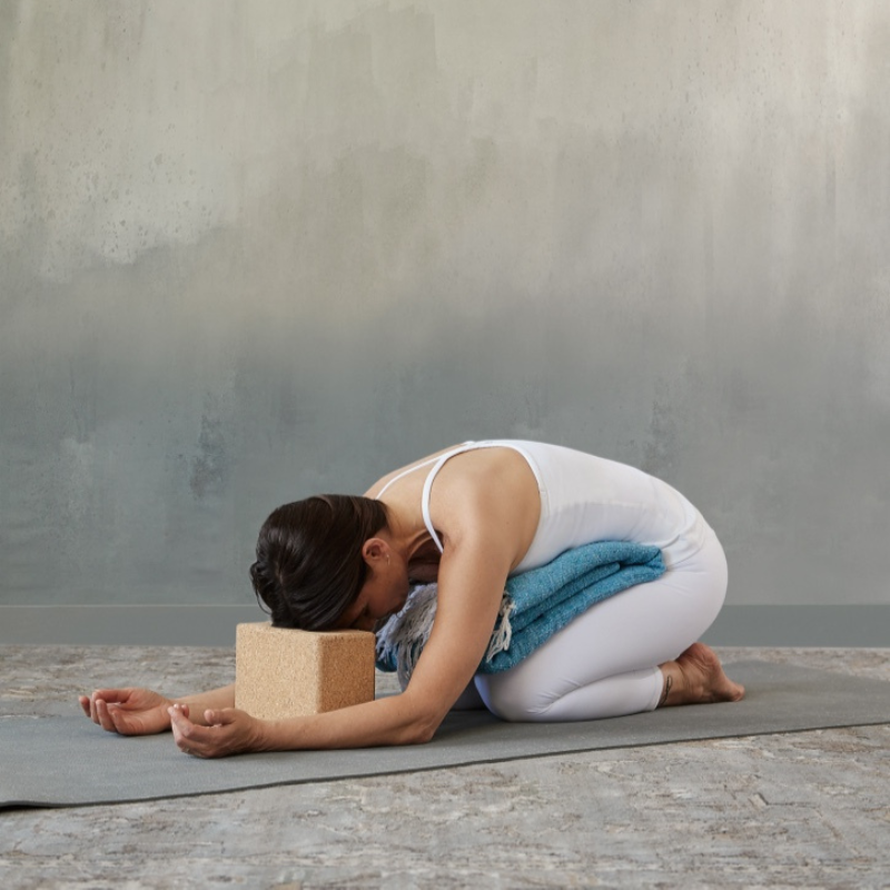 A woman practicing restorative yoga with minimal props indoors, in a kneeling forward bend position on a yoga mat, with a yoga block supporting her forehead and holding a rolled towel.