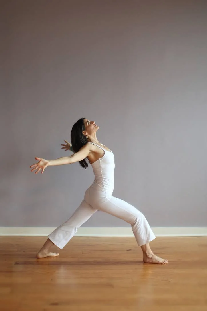 Woman practicing yoga in a lunge position inside a room, wearing workout clothes and a headset microphone, with a dog sleeping in a bed nearby.