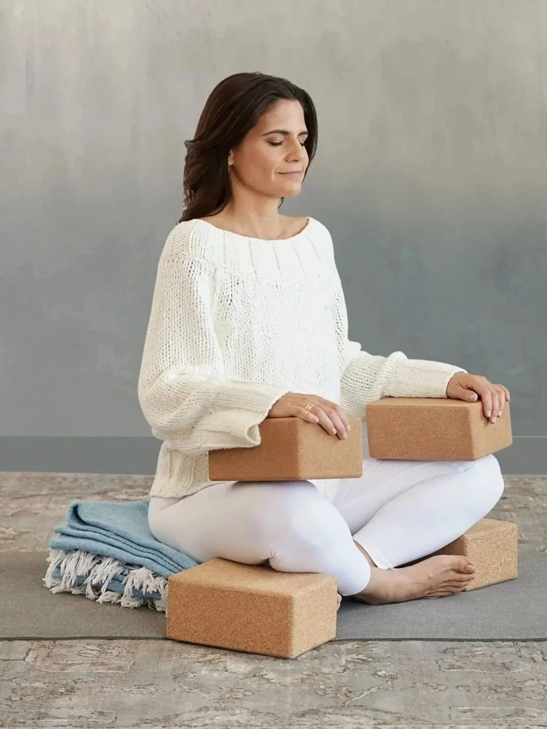 A woman sitting cross-legged on a yoga mat, holding yoga blocks, with a calm and focused expression, practicing meditation.