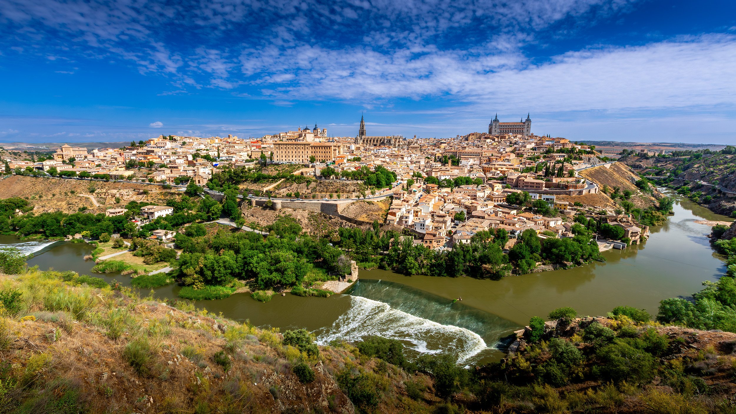 A panoramic view of Toledo Spain with the Tagus river flowing in the foreground and a blue sky with scattered clouds.