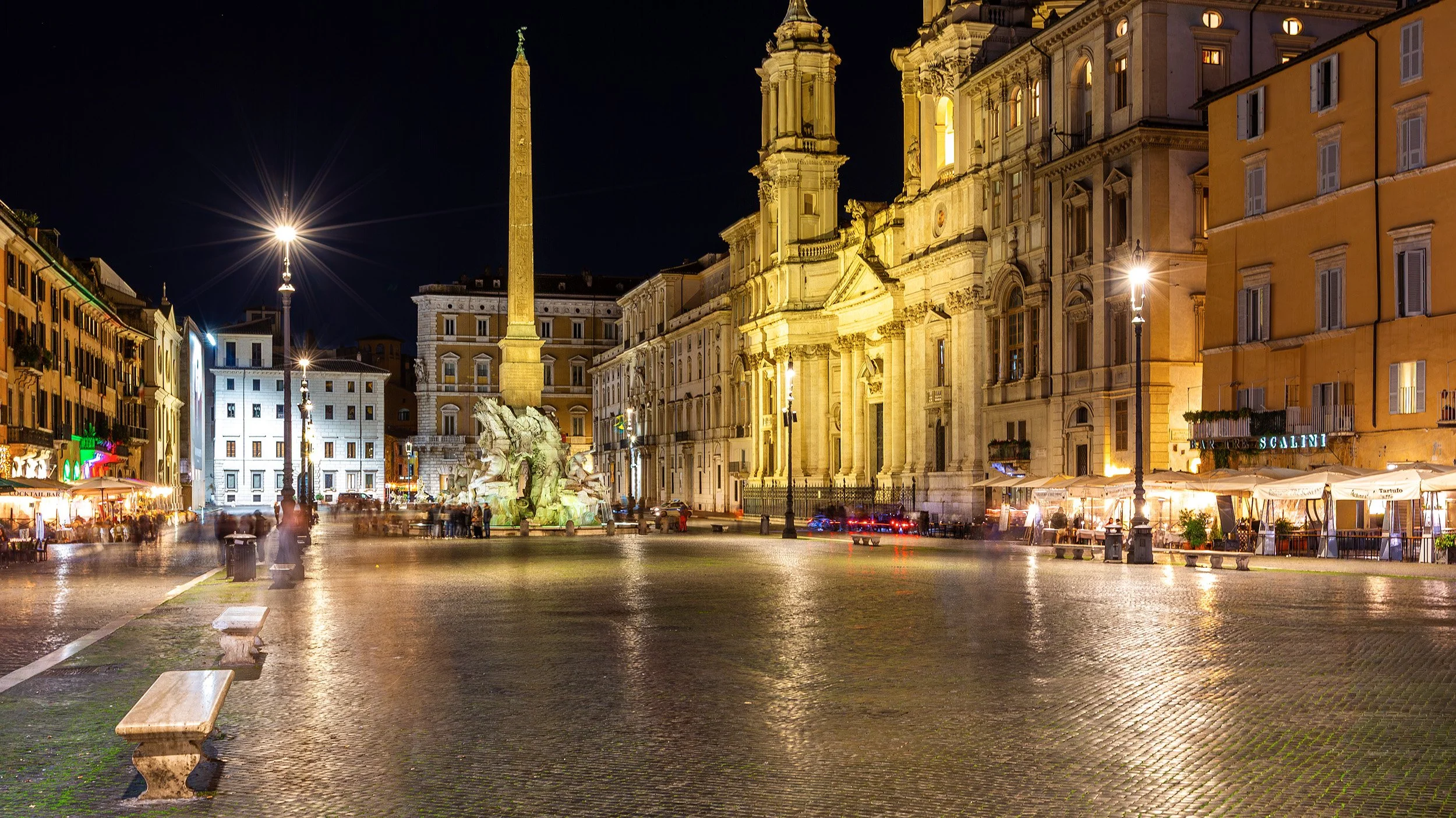 Night view of Piazza Navona in Rome, Italy, with illuminated Baroque buildings, a fountain, and street lights, with people walking and outdoor cafes.
