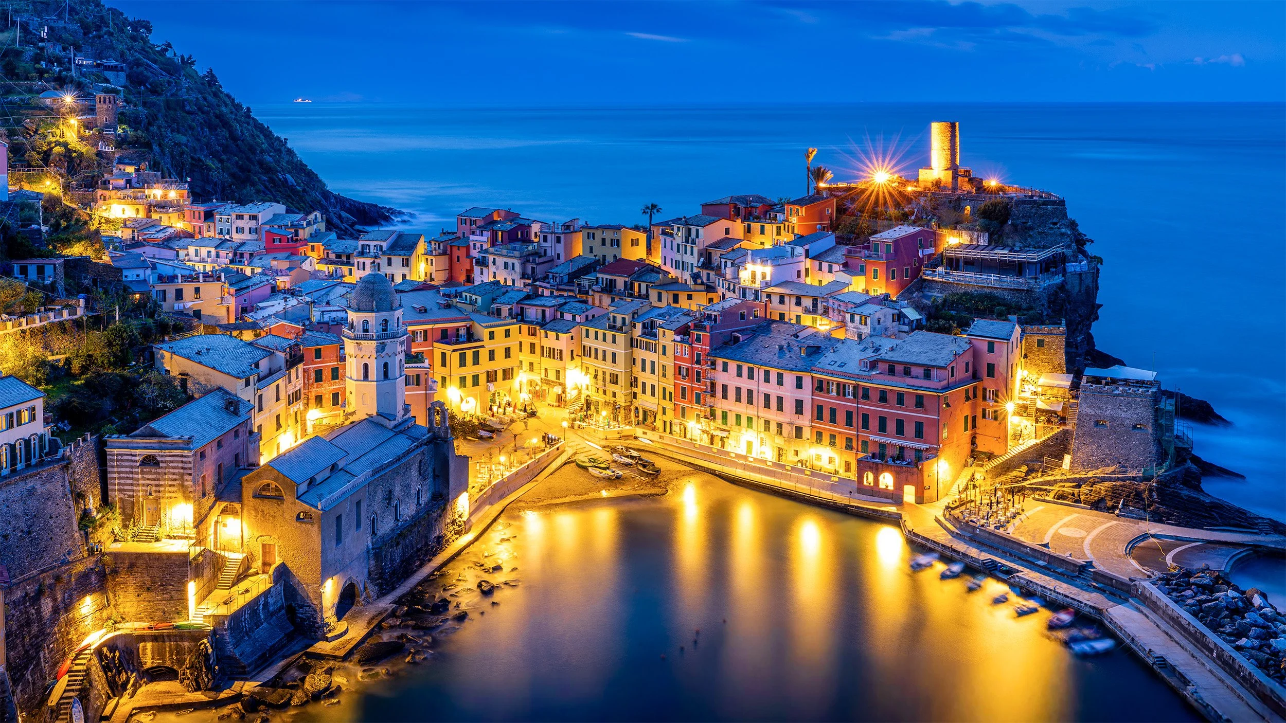 The colourful seaside village of Vernazza at twilight with illuminated buildings, a harbour, boats, and a hill with houses and a tower, overlooking the ocean.