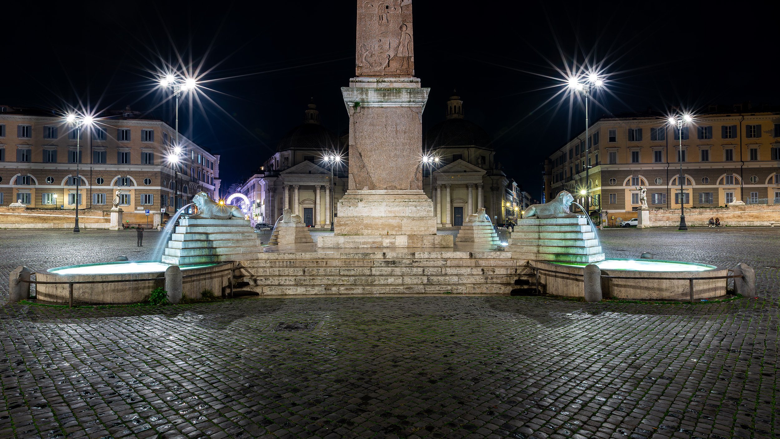 Night view of a Piazza del Popolo with a 3,200 year-old Egyptian obelisk, lion fountain statues with water, surrounding by twin Baroque churches (Santa Maria dei Miracoli and Santa Maria in Montesanto), and bright streetlights.