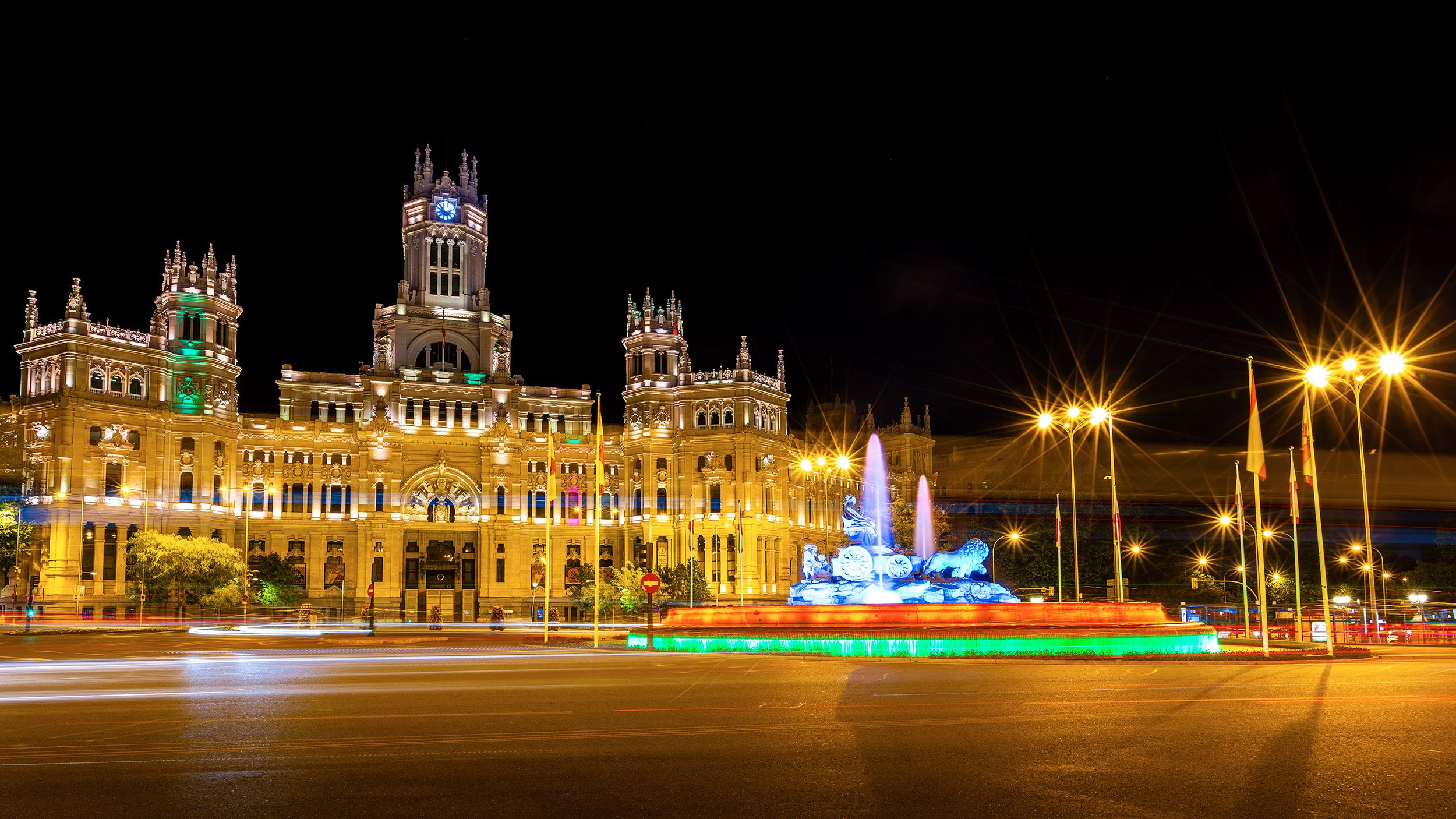 Night view of Palacio de Cibeles (sede del Ayuntamiento de Madrid) with illuminated towers, a clock, and a fountain in front, with colorful lights and light trails from moving vehicles.