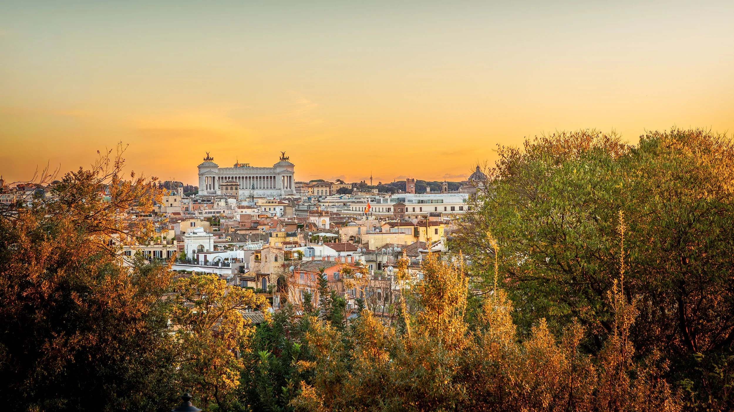 View of Rome at sunset with the Monument to Victor Emmanuel II in the background and trees in the foreground.