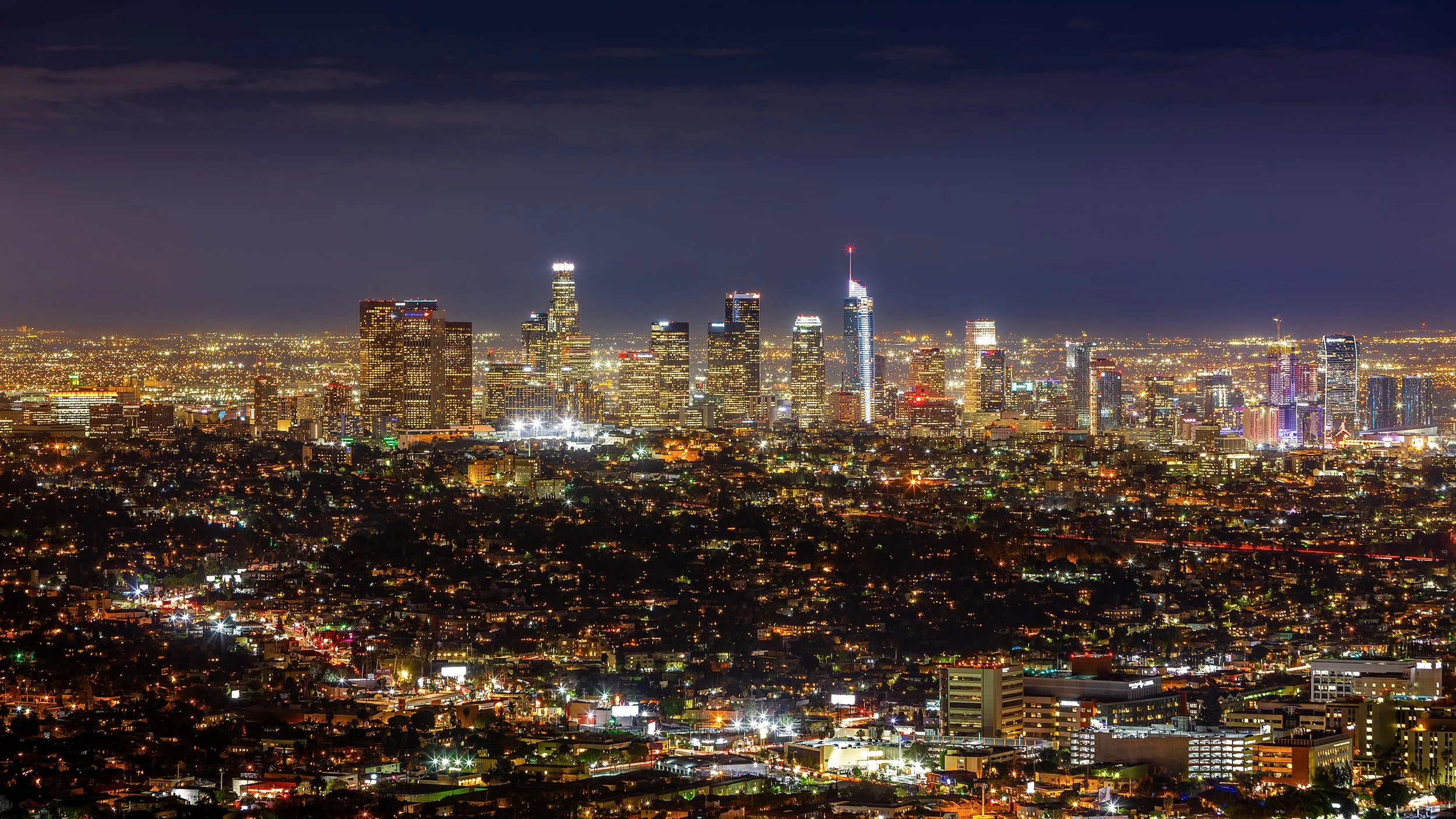 Nighttime view of the downtown Los Angeles skyline with illuminated skyscrapers and city lights.