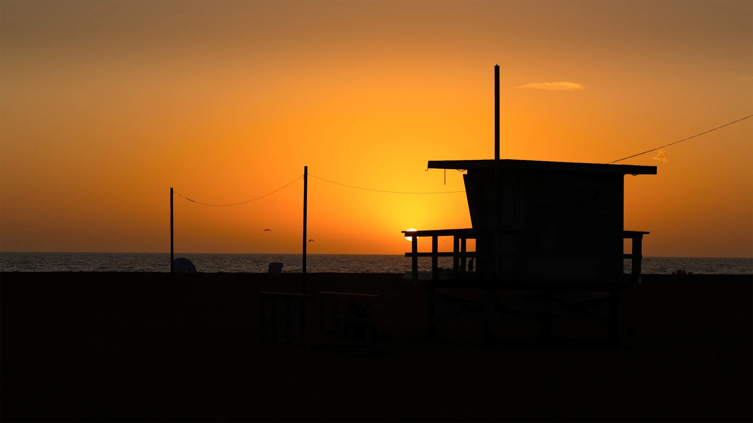 Silhouette of a lifeguard tower on the beach during sunset, with the sun just above the horizon and a few clouds in the sky, calm ocean, and a few birds flying.