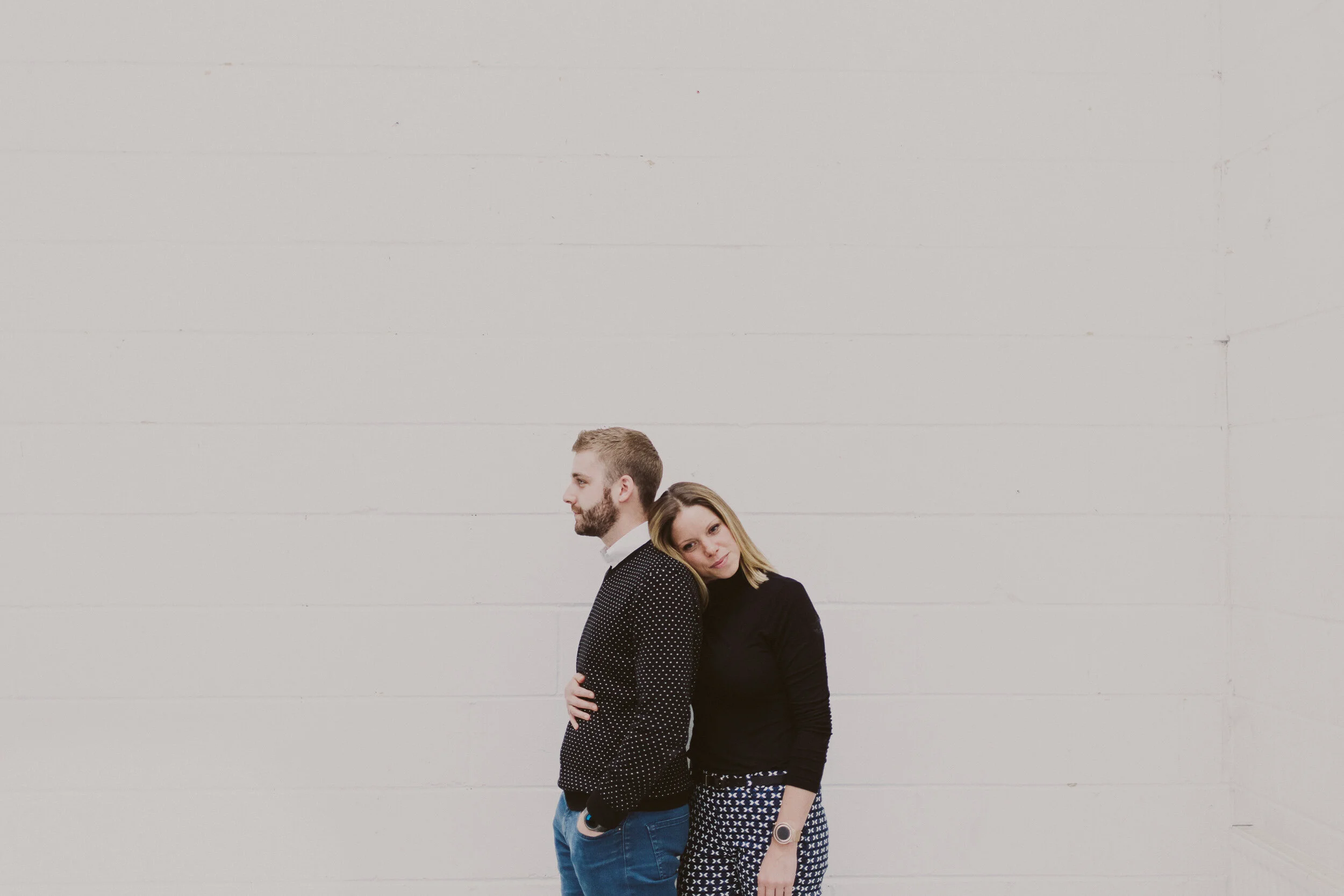 A woman and a man standing back-to-back against a plain white wall. The woman is resting her head on the man's shoulder and has a gentle smile. The man has his hands in his pockets and is looking away. Both are dressed casually.
