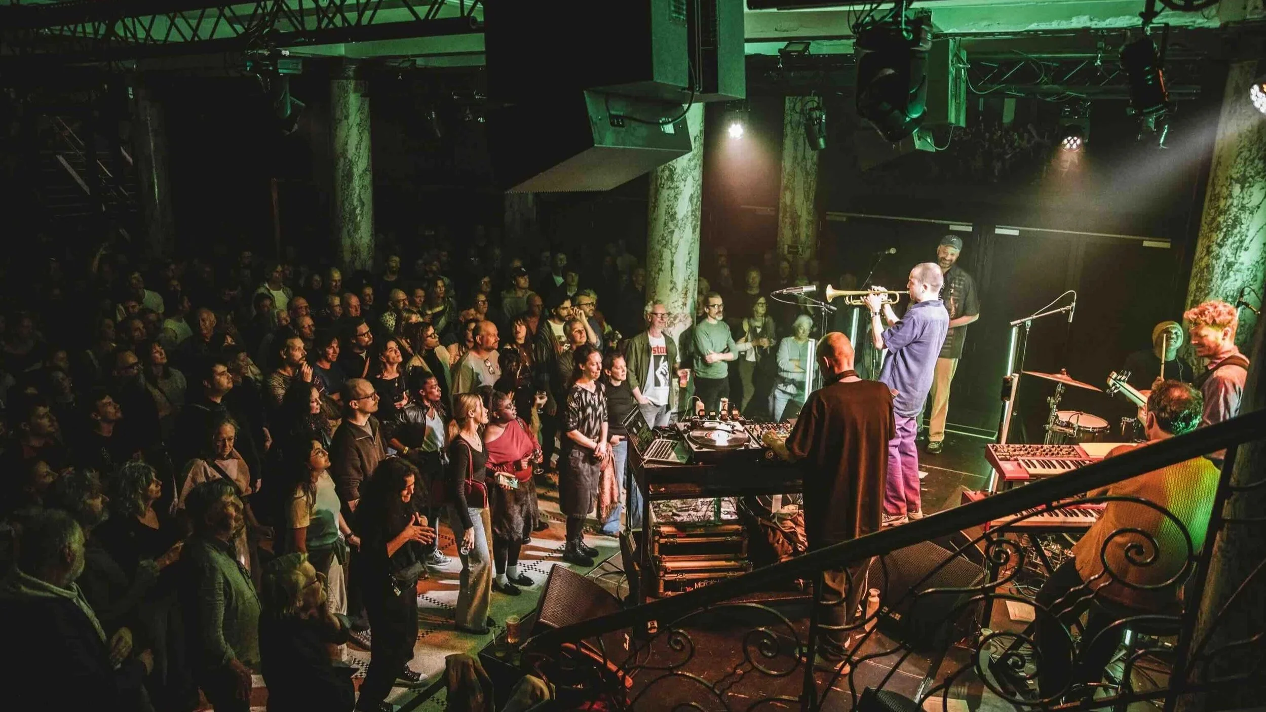 A live music performance at an indoor venue featuring a band on stage with a diverse audience watching attentively, illuminated by stage lighting with exposed beams and green marble pillars in the background.