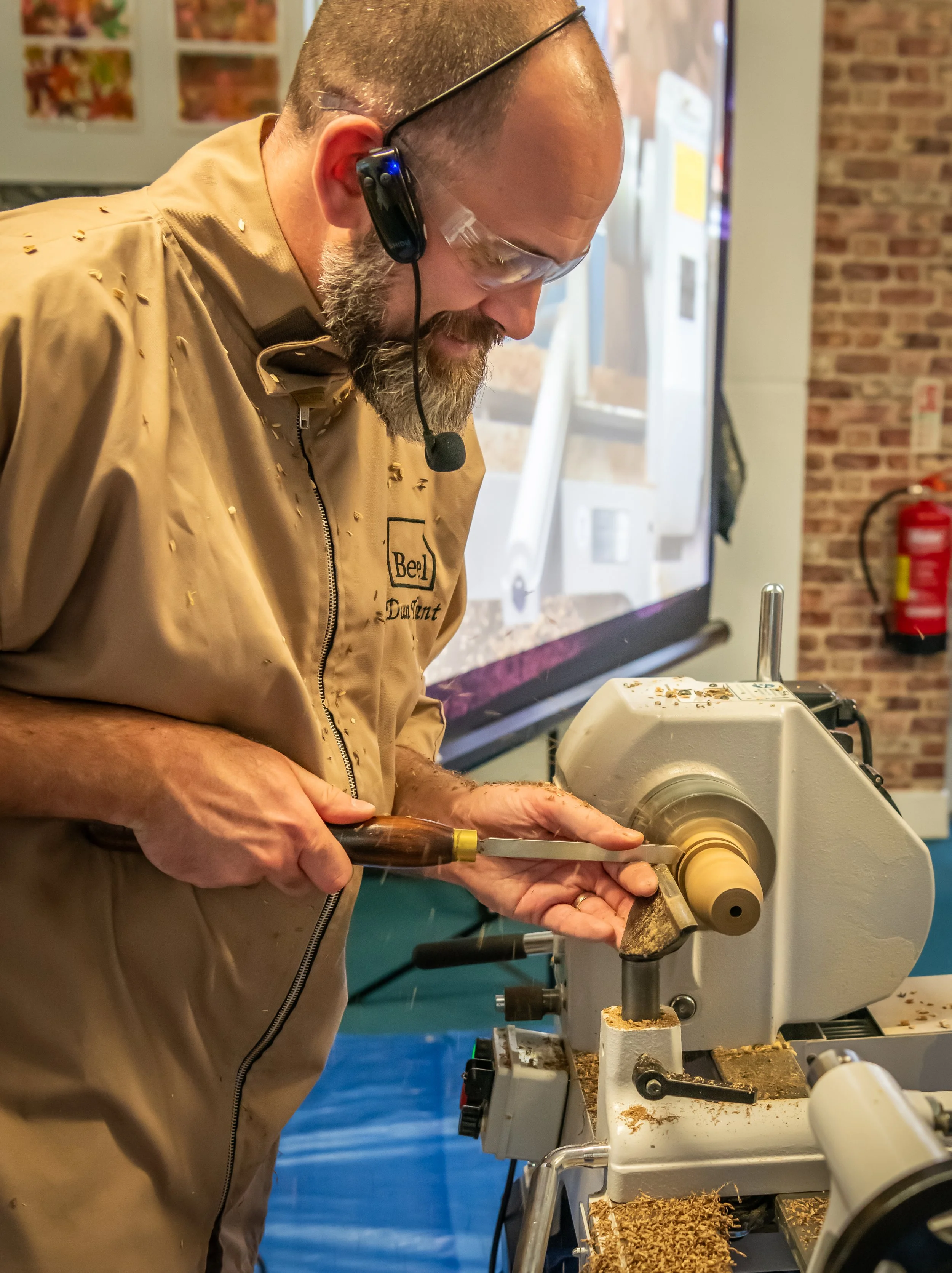 Man wearing safety glasses and a tan jacket operating a wood lathe, shaping a piece of wood in a woodworking shop.