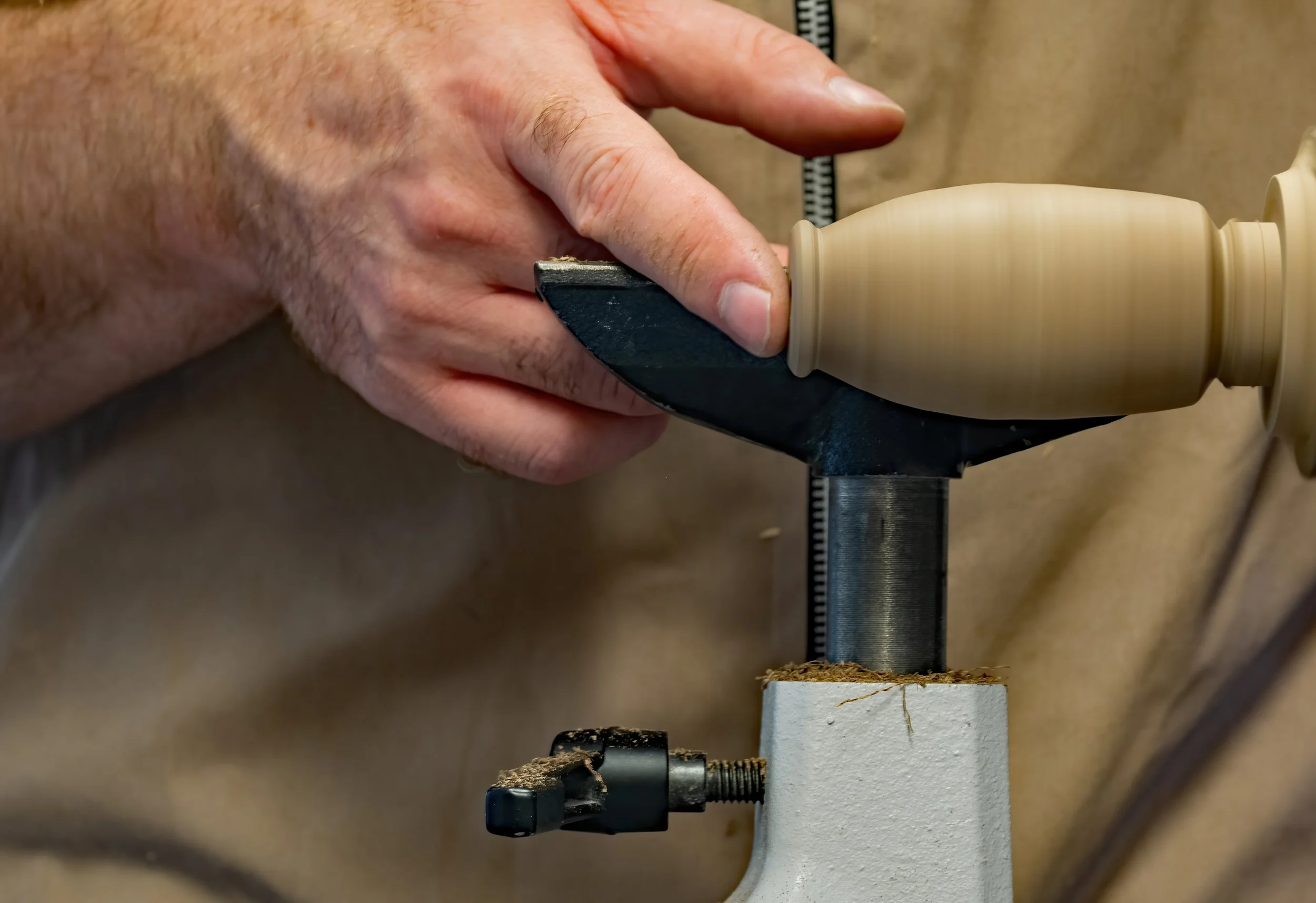 Close-up of a person's hand holding a chisel, working on a piece of wood mounted on a lathe machine in a woodworking shop.