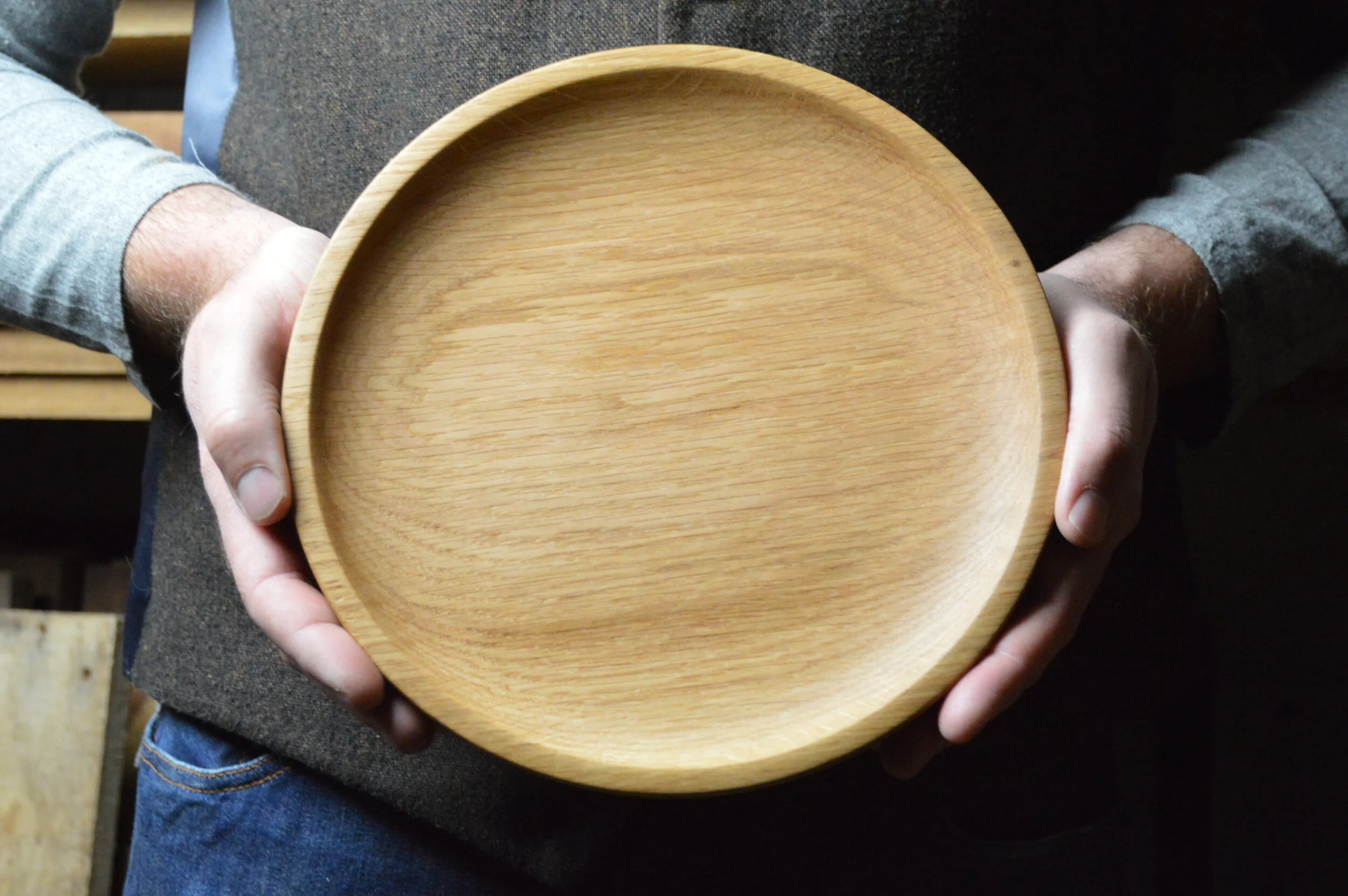 A person holding a round wooden tray with a smooth finish, with their hands visible on either side.