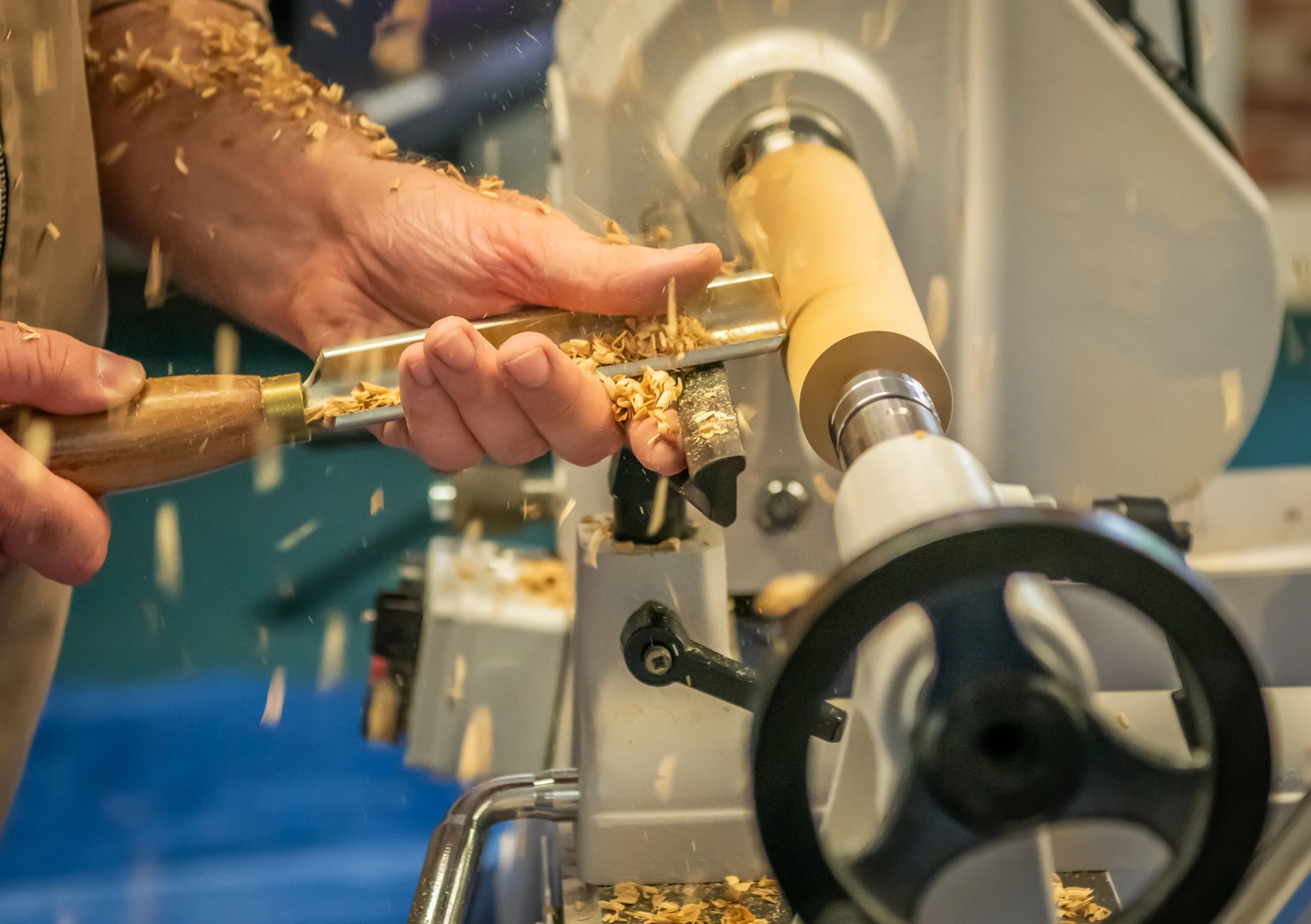 A person operating a woodworking lathe machine shaping a piece of wood, with wood shavings flying around.