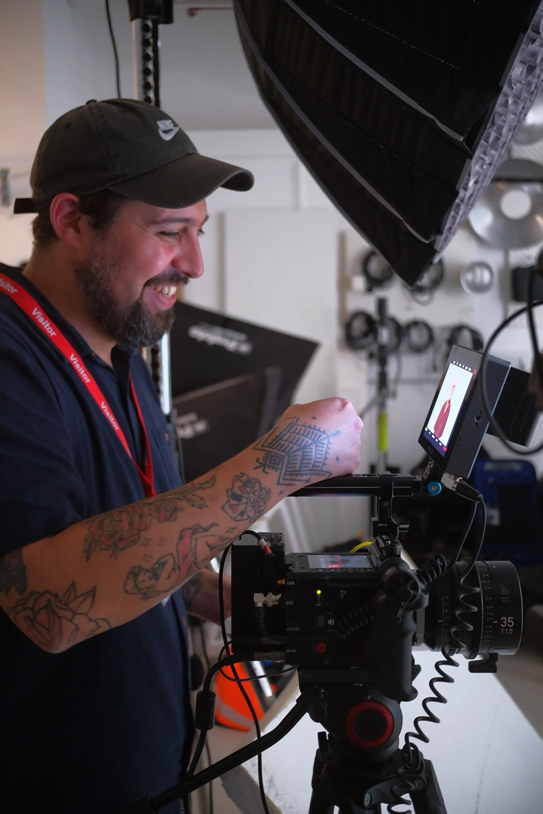A man with tattoos on his left arm, wearing a black Nike cap and a black shirt with a red lanyard, is smiling while looking at a monitor attached to a professional camera setup. The camera is positioned on a tripod and there is sort of LED or light attached to it. The background shows a wall with some equipment mounted on it and a large black softbox or diffuser.