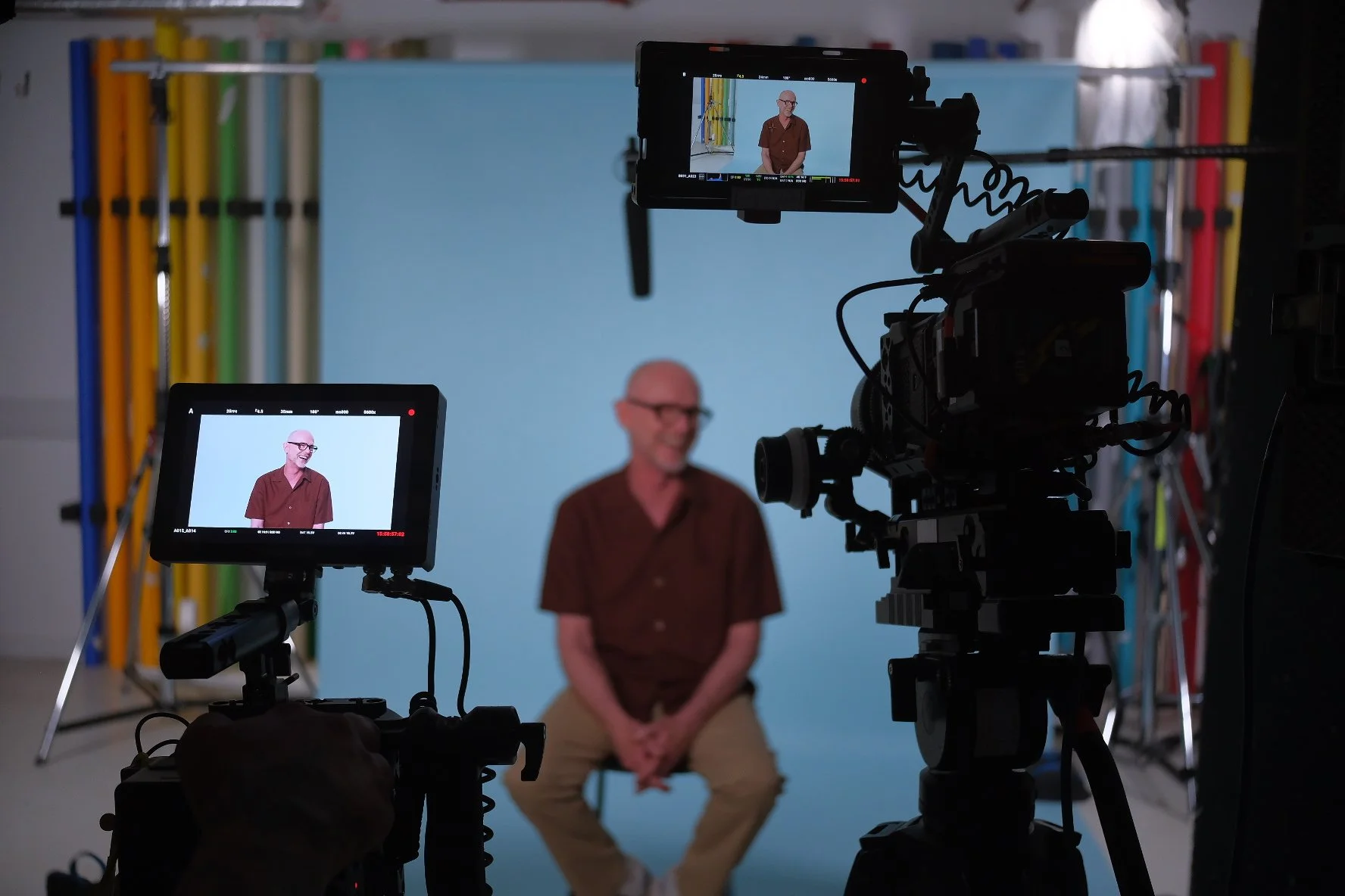 A man seated in front of a blue screen during a video shoot, with cameras and monitors recording him.
