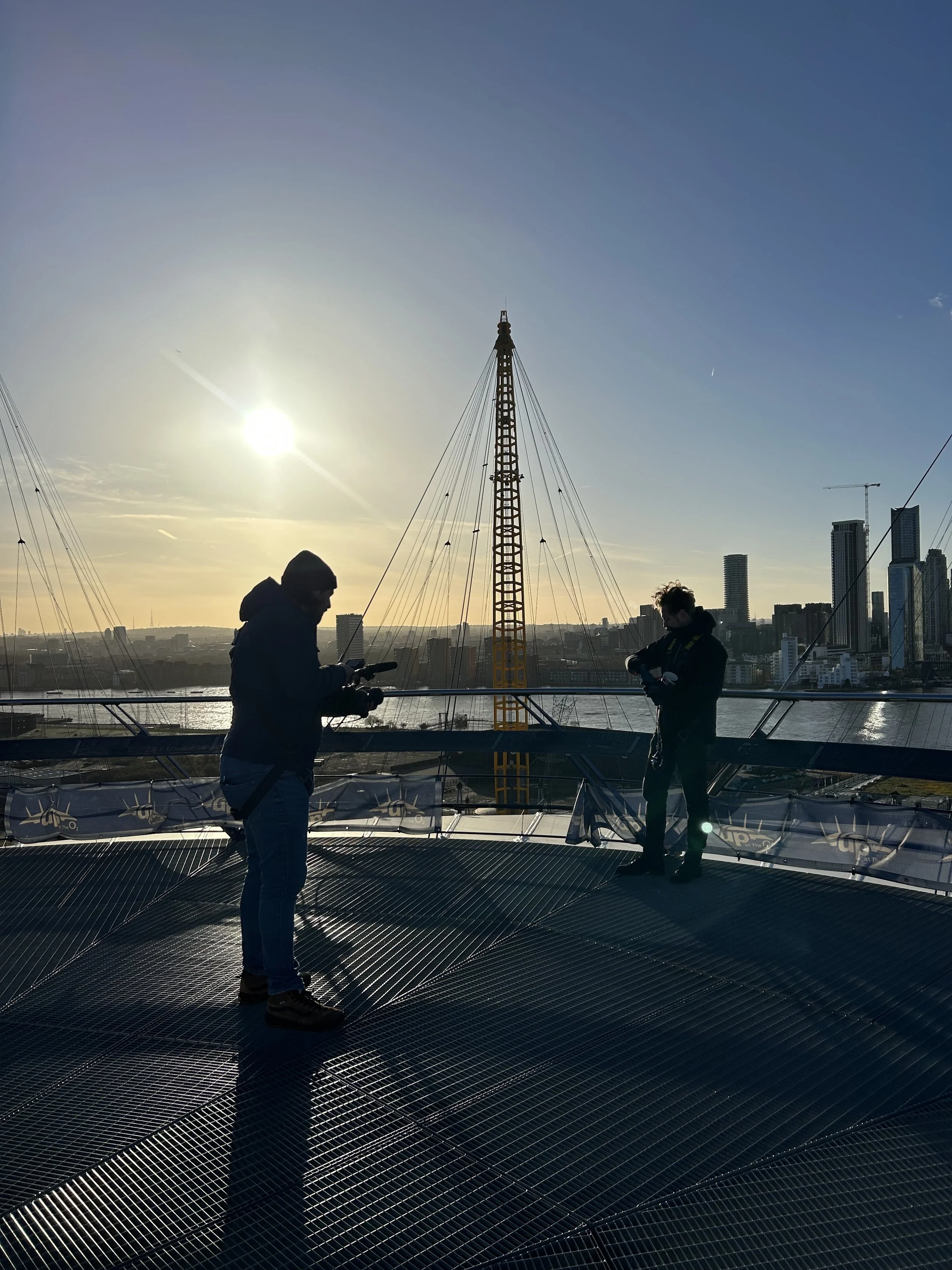 Silhouettes of two people standing on a rooftop or observation deck at sunset, with a city skyline and tall buildings in the background.