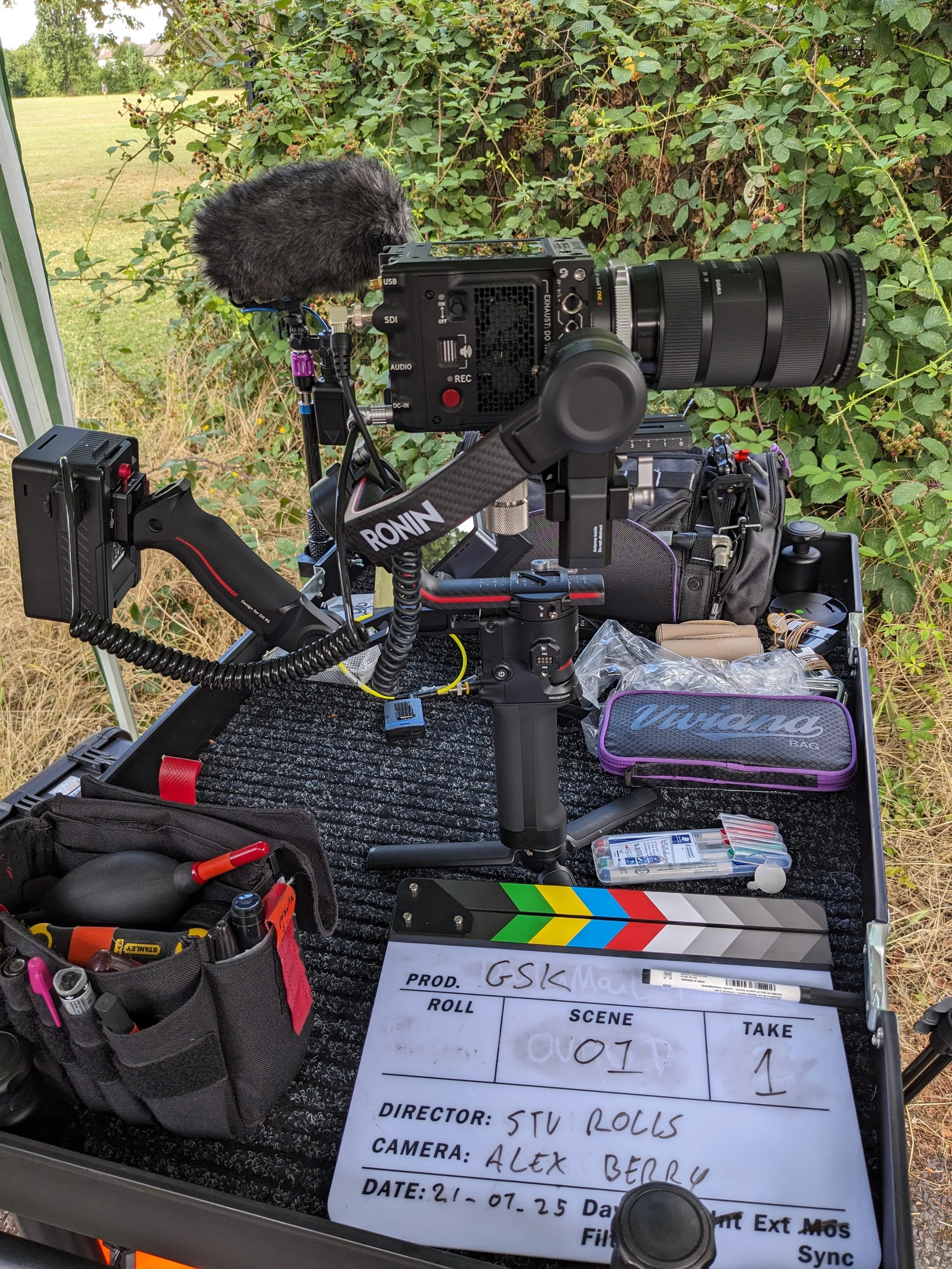 A professional camera setup on a tripod, with various accessories, on a table outdoors. Behind is greenery and an open field.