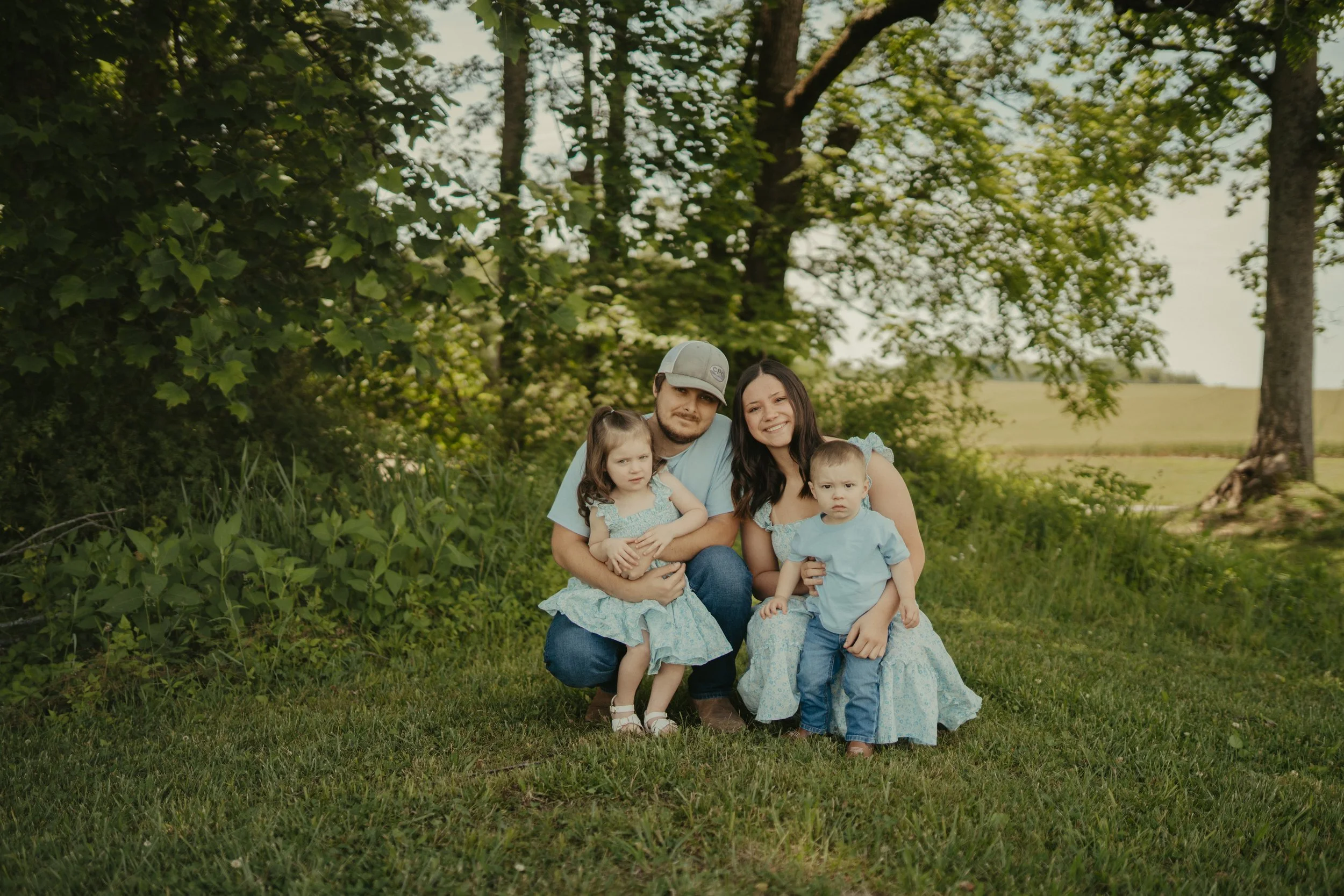 A family of five sitting on grass in a park with trees in the background. The father, mother, and three young children are smiling and looking at the camera.