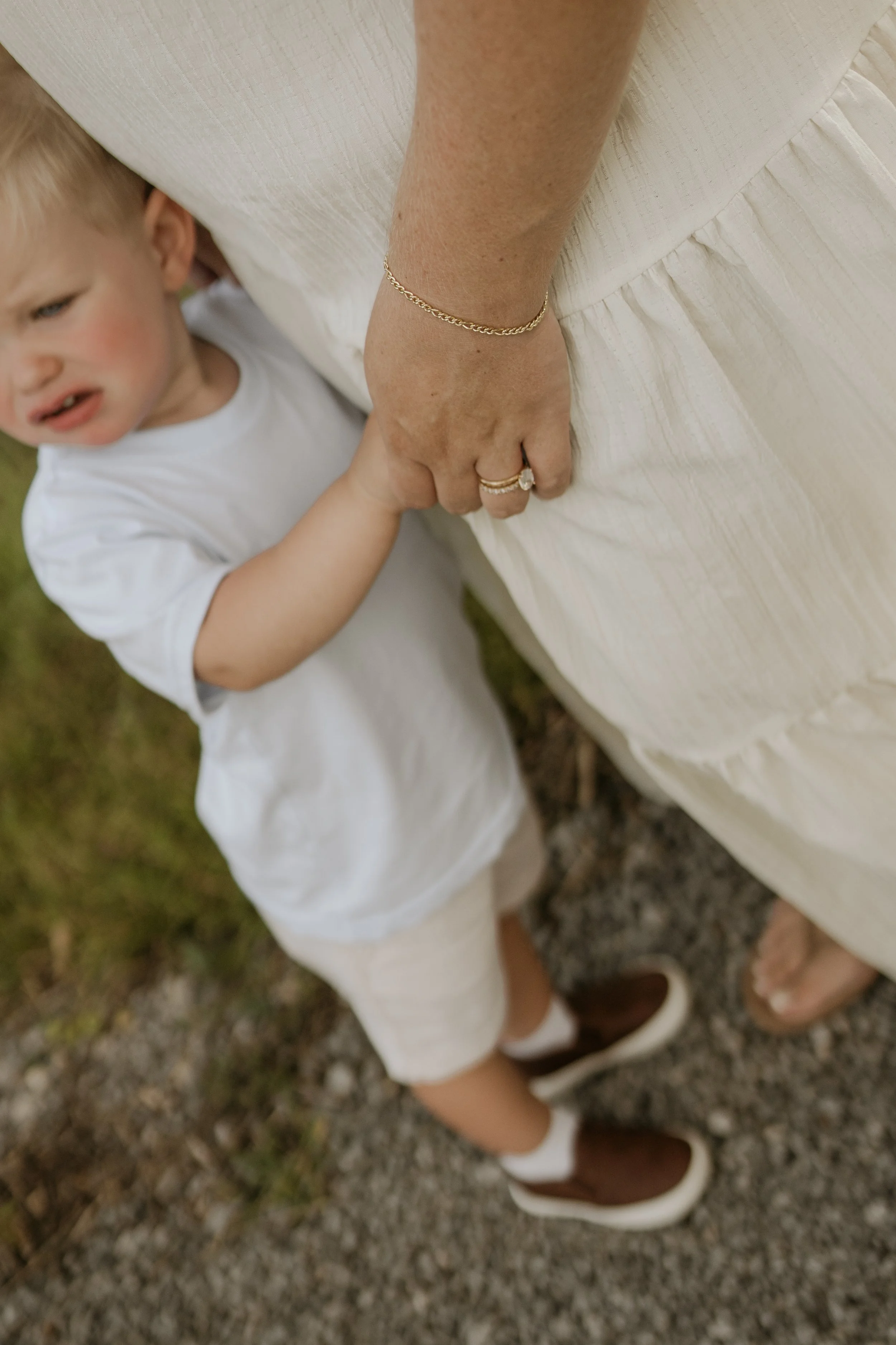 A young child holding a woman's hand outdoors on a gravel surface. The woman is wearing a beige dress, jewelry, and white shoes, while the child is dressed in a white outfit and brown shoes.