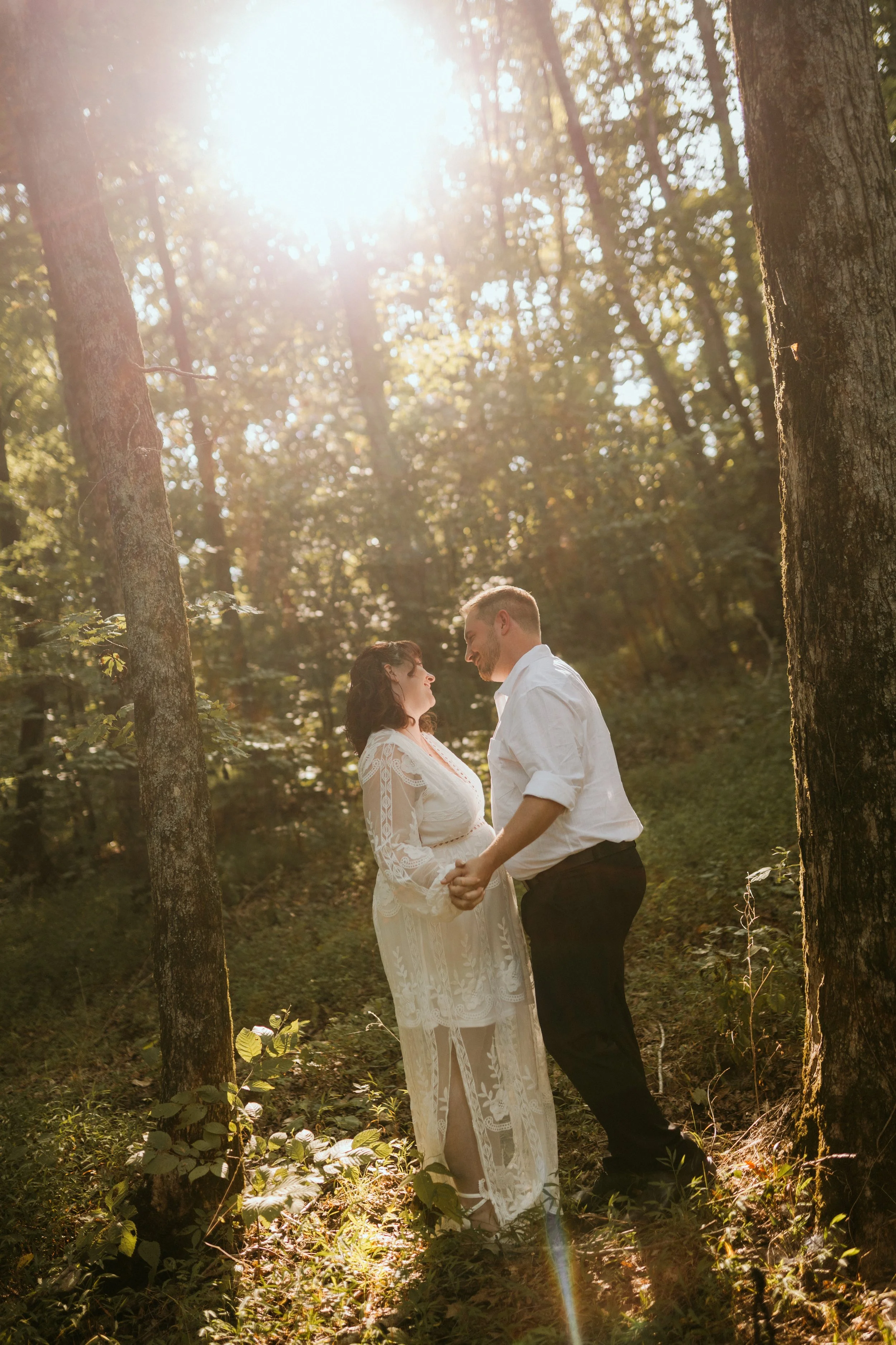 A couple in wedding attire holding hands and looking at each other in a sunlit forest during daylight.