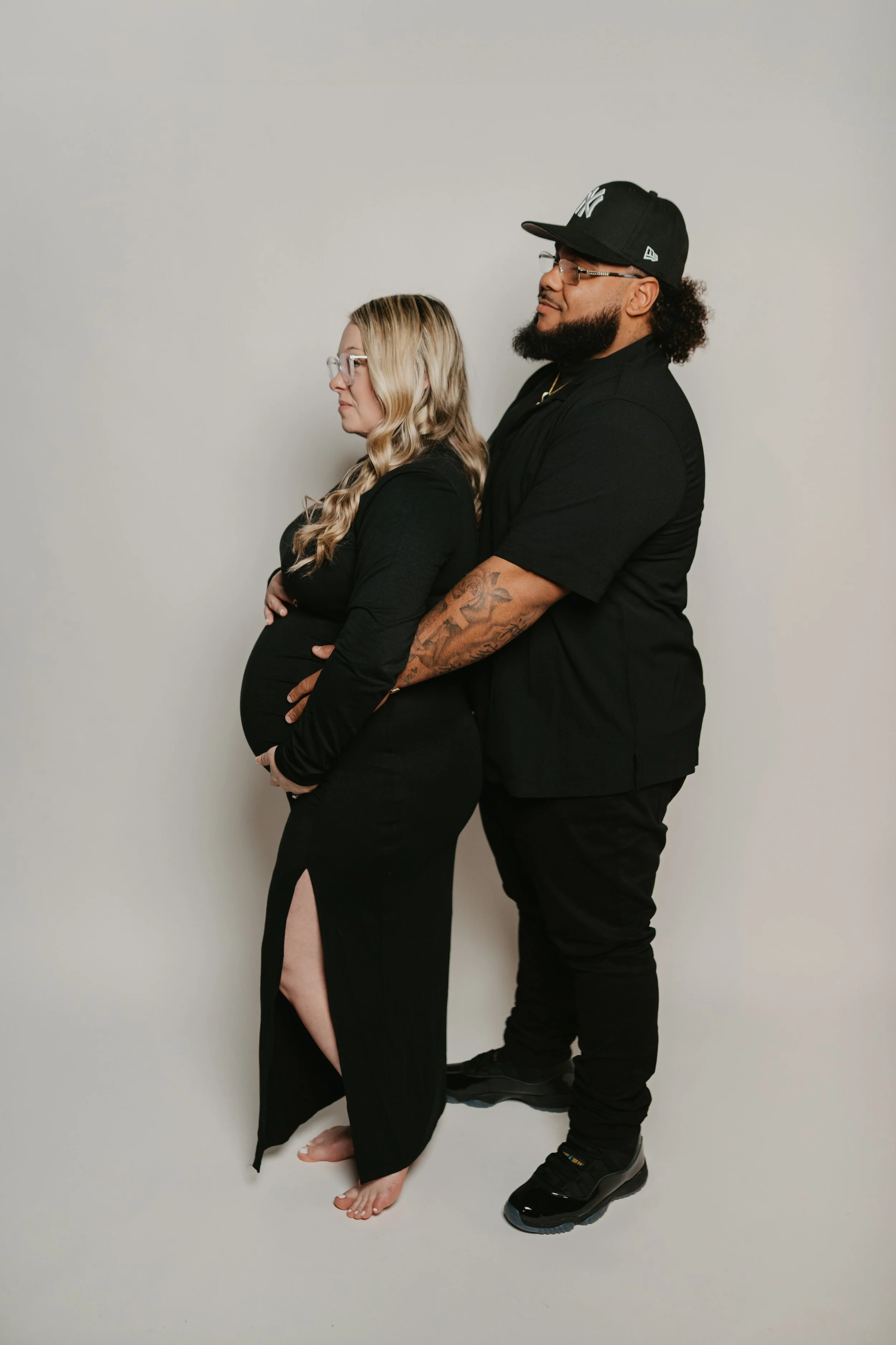 A pregnant woman in a black dress with a slit revealing her leg, standing barefoot, with blonde wavy hair and glasses, while a man in black with a cap and glasses, sporting tattoos and black sneakers, stands behind her, both leaning against a plain l