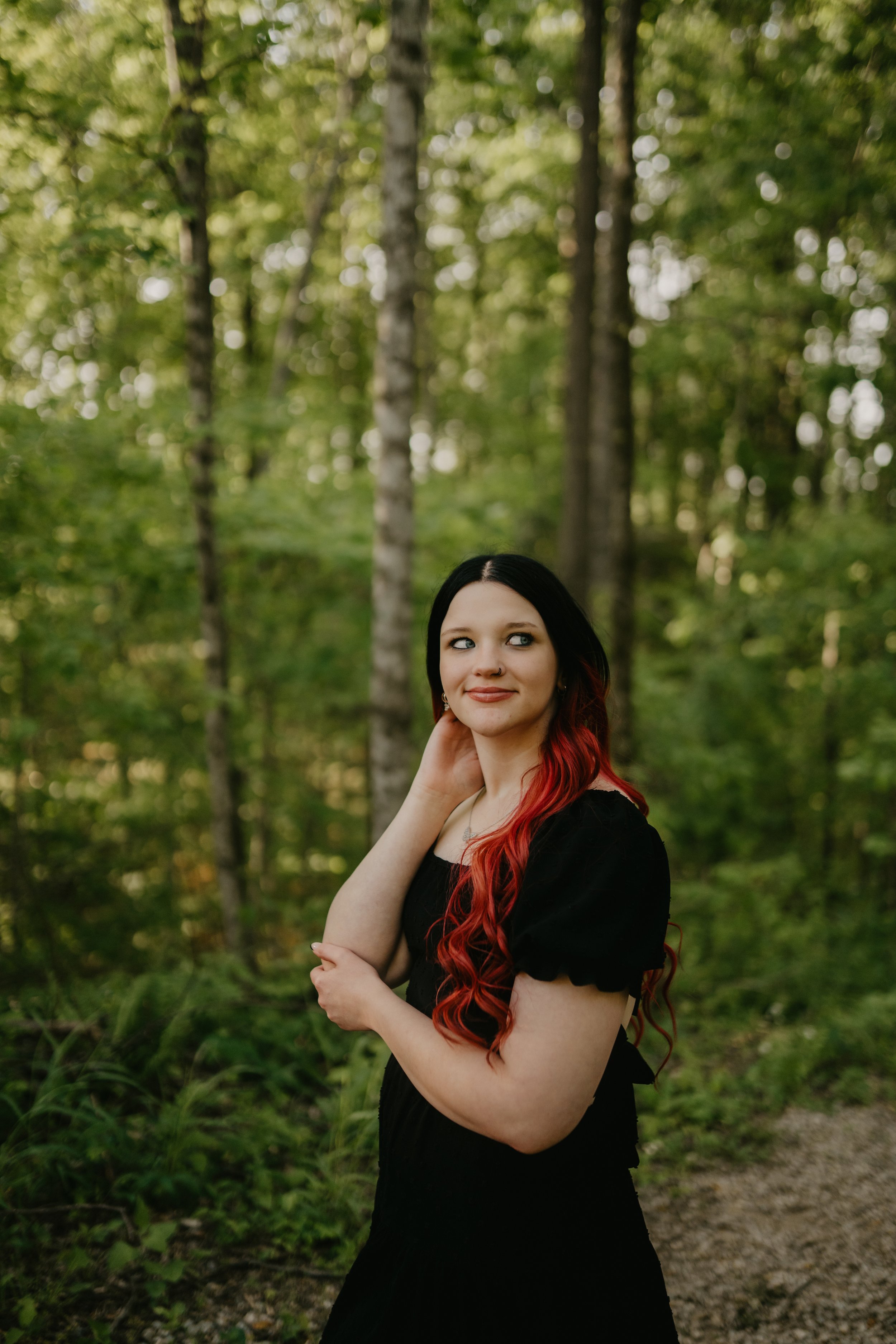 A woman with long black and red hair, wearing a black dress, standing in a forested area with green trees and foliage in the background, smiling softly.