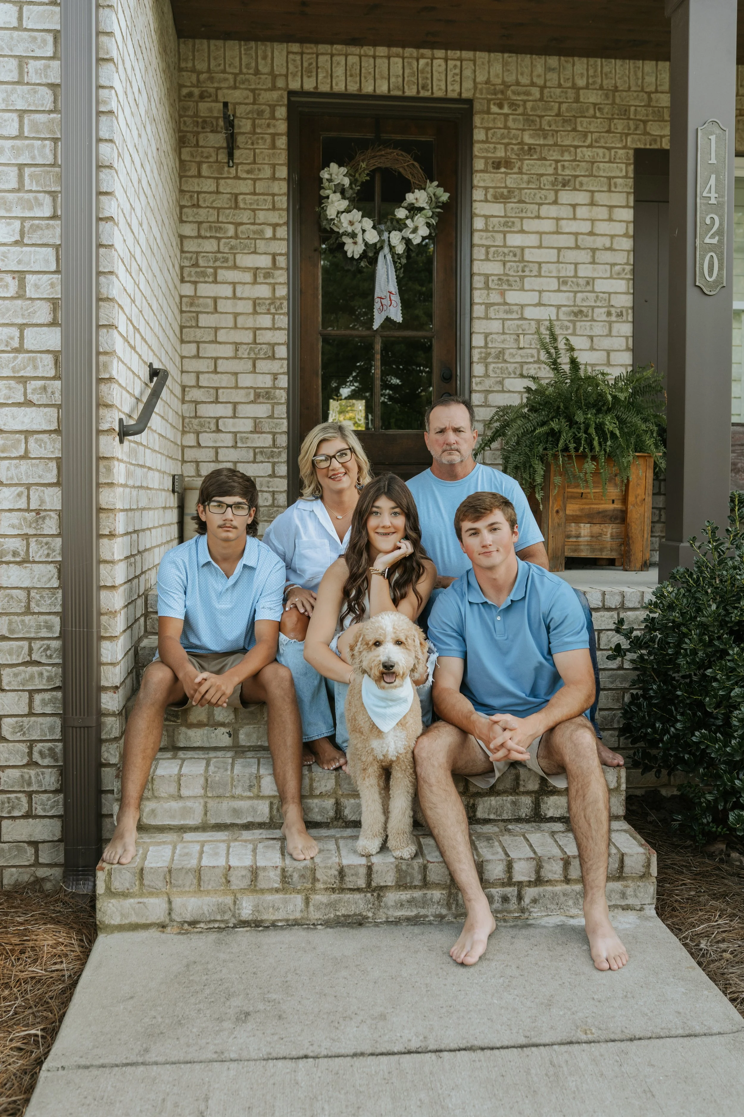 A family of five with three males, one female, and a dog, all sitting on the front steps of a house. The family is dressed casually with all males in blue shirts, and the woman in a white top. The dog, a goldendoodle, is in front of the woman. The ho