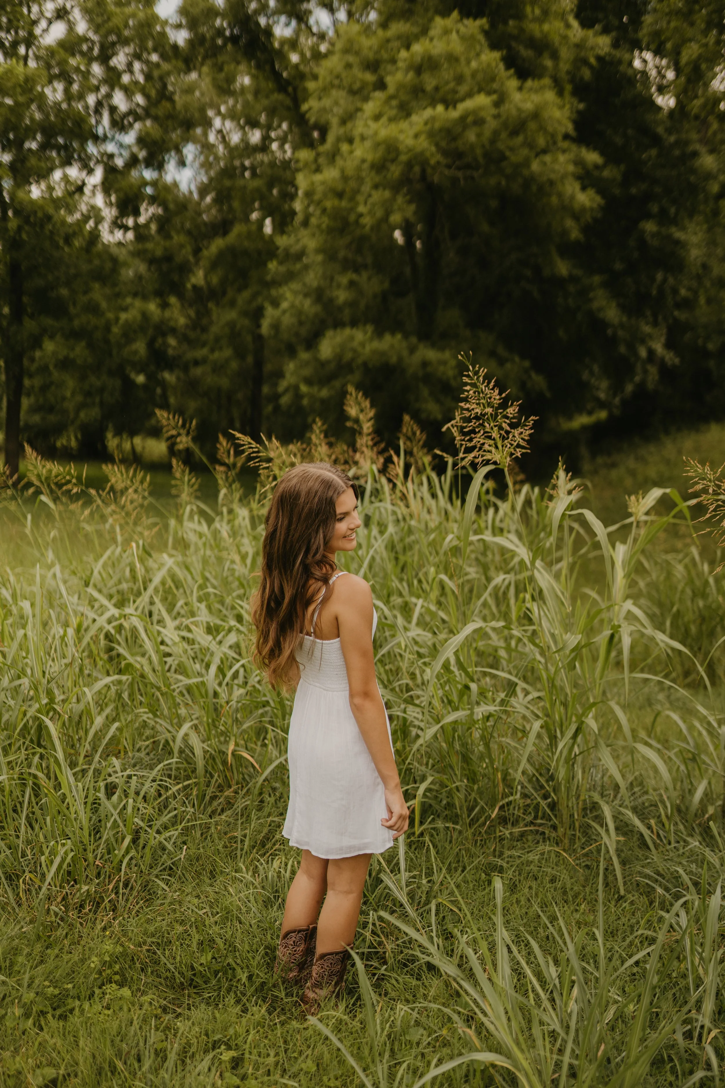 A young woman with long brown hair wearing a white dress and cowboy boots standing in a grassy field with tall grass, trees in the background, and a cloudy sky.